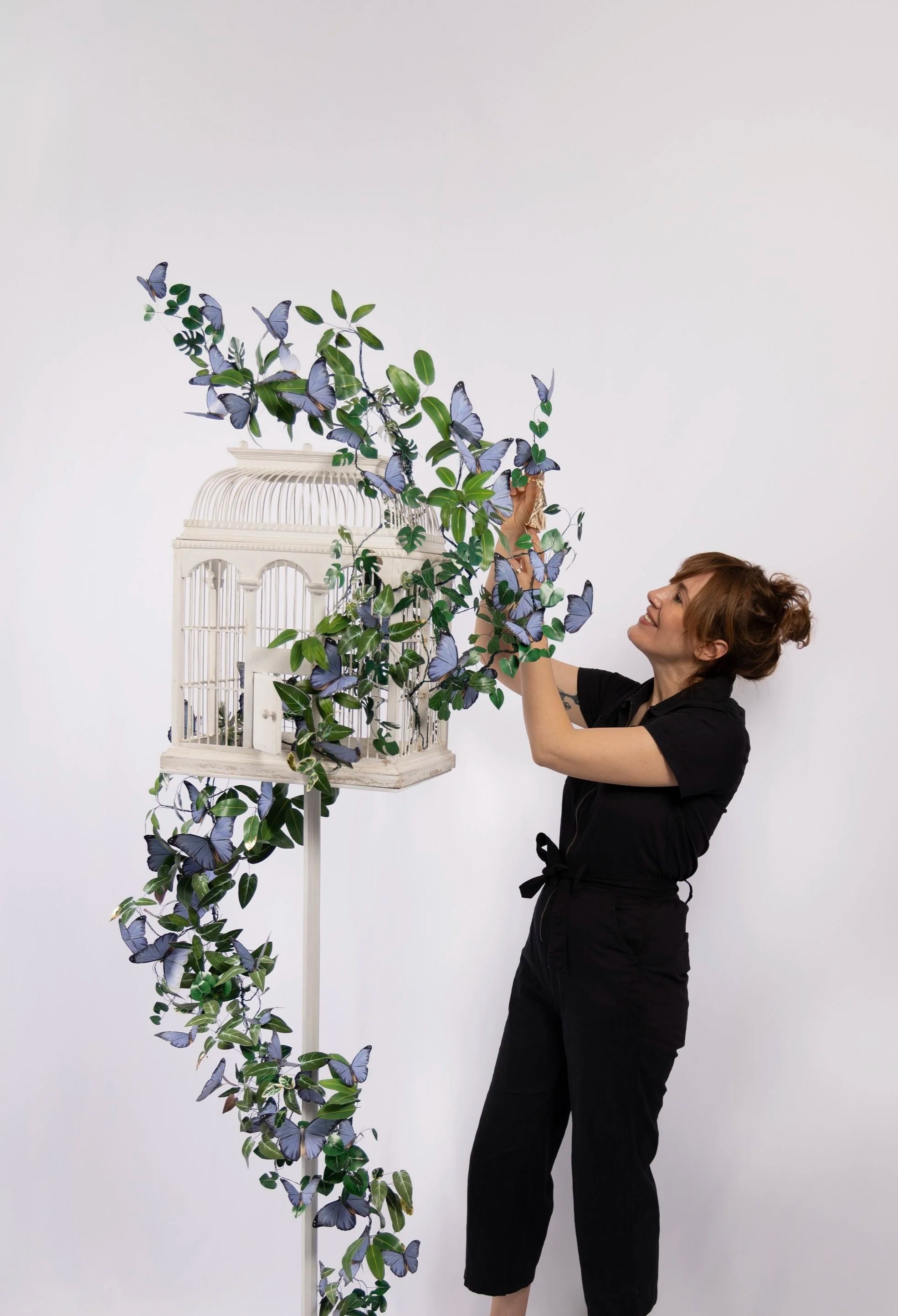 Clare Celeste Börsch arranging a vine of paper leaves and blue butterflies on a vintage birdcage in her studio