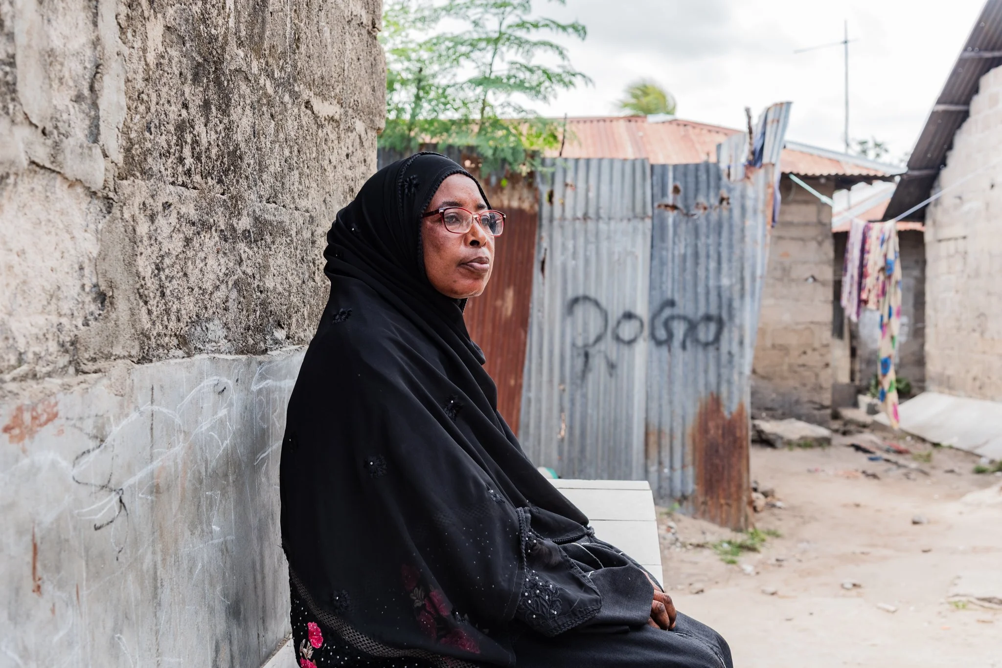 An older woman wearing glasses and a black headscarf sits on a bench against a concrete wall in Zanzibar, Tanzania.