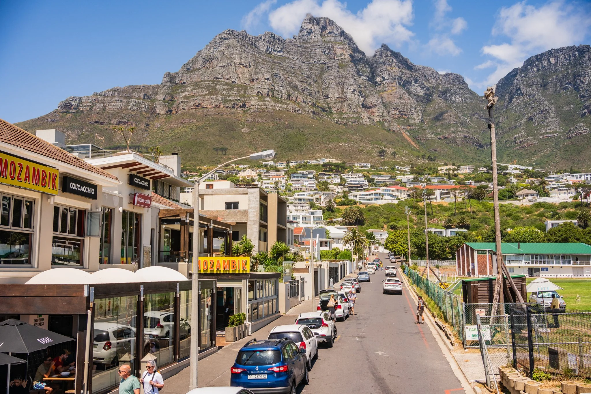 Street view in a hilly area with commercial buildings, parked cars, pedestrians, and a mountain in the background.