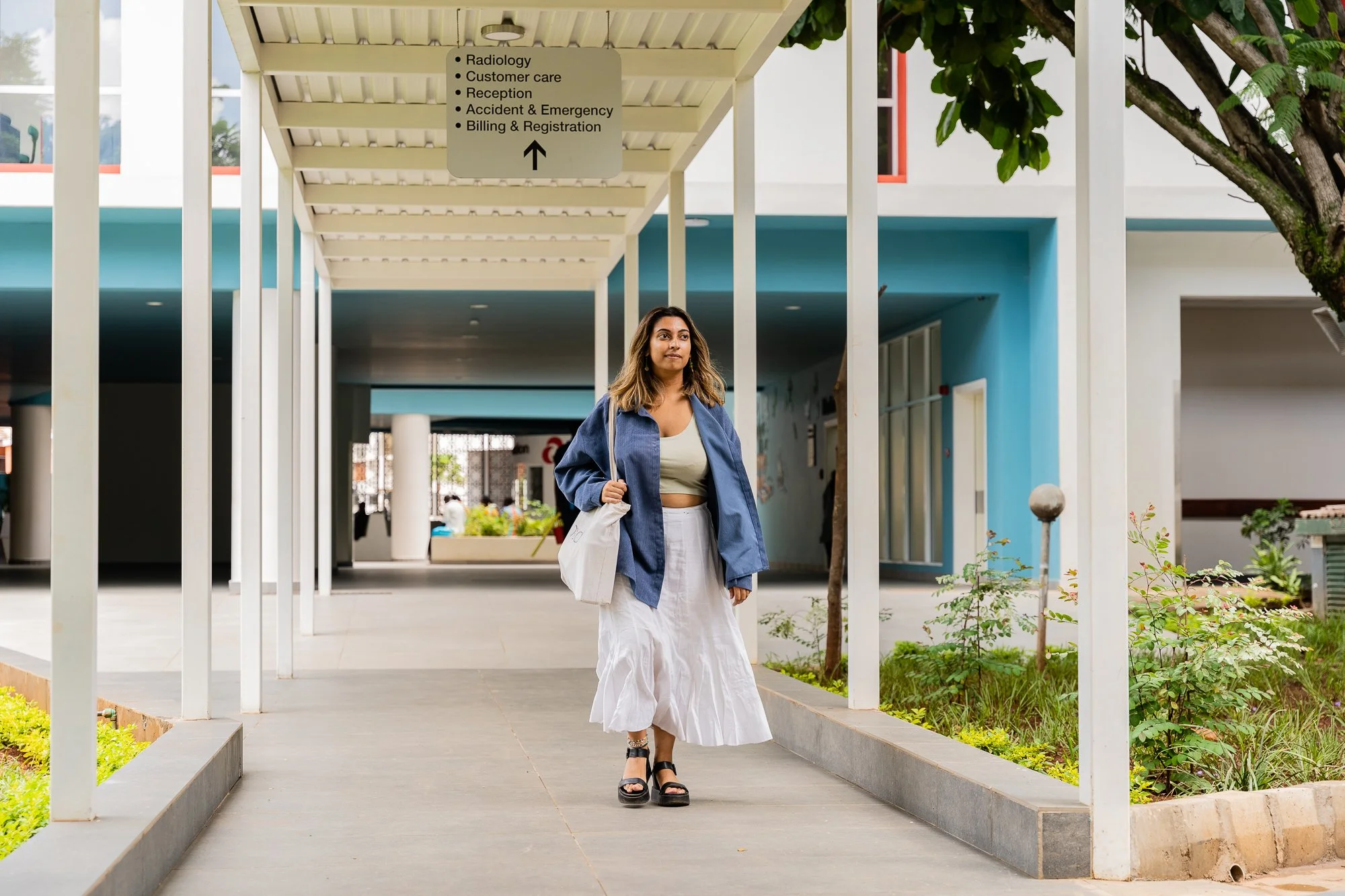 AKFC International Youth Fellowship fellow walking under a covered walkway at Aga Khan Hospital in Kisumu, Kenya, carrying a white tote bag with directional signage for hospital departments visible overhead.