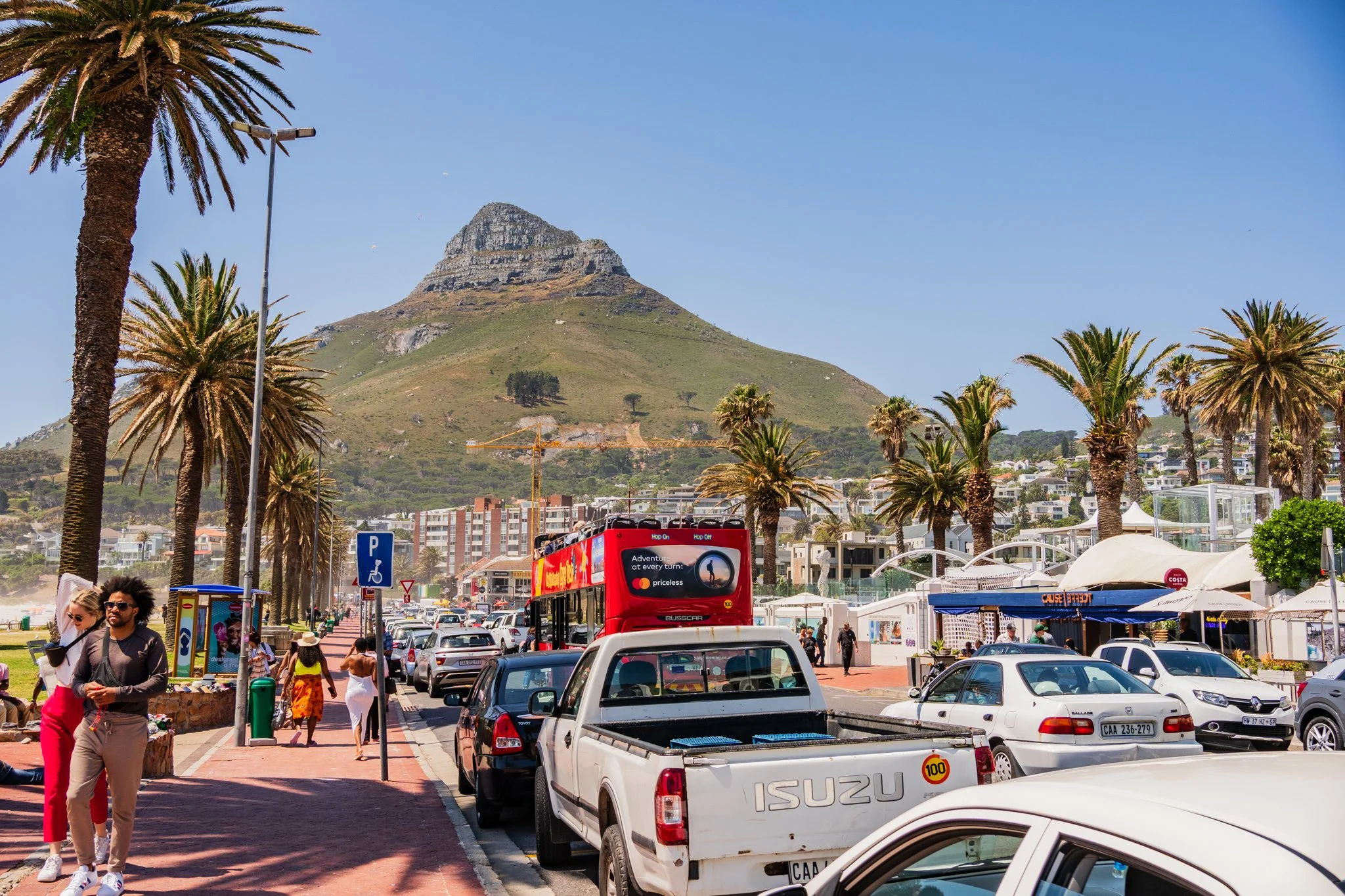 Beachside street with palm trees, parked cars, and people walking, with a mountain in the background under a clear blue sky.