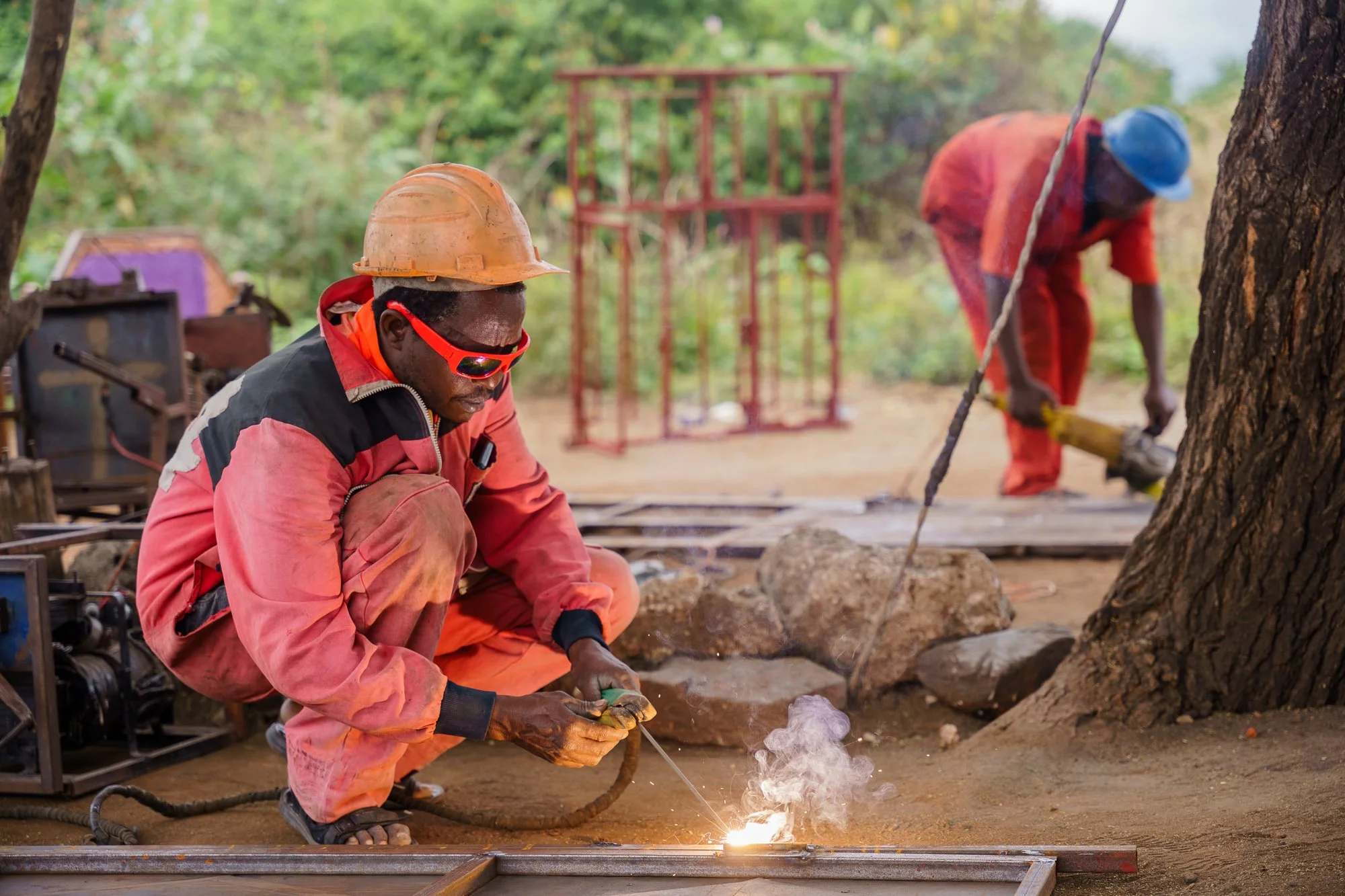 Two workers welding and using tools outdoors near a tree in Babati, Manyara, Tanzania.