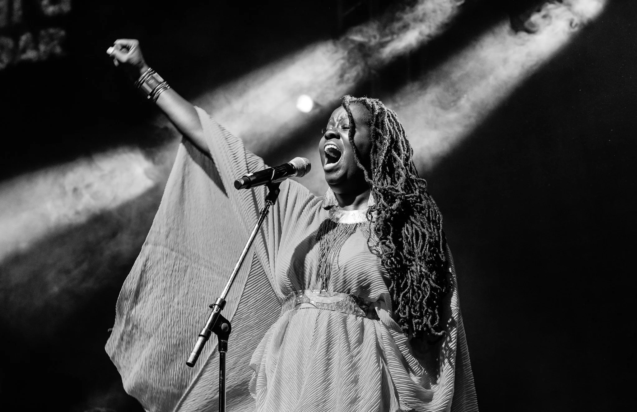 A black and white photograph of a woman singing passionately into a microphone, with her right arm raised and her mouth wide open, performing on stage with dramatic lighting in Zanzibar at the Sauti za Busara Music Festival in 2018.