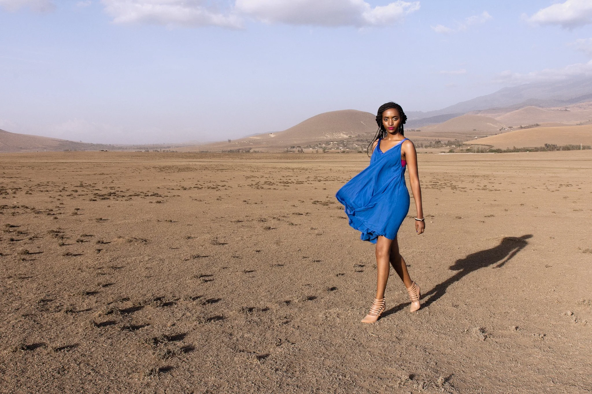 A woman in a blue dress walking across a dry, desert landscape with mountains in the background and a partly cloudy sky in Arusha, Tanzania.