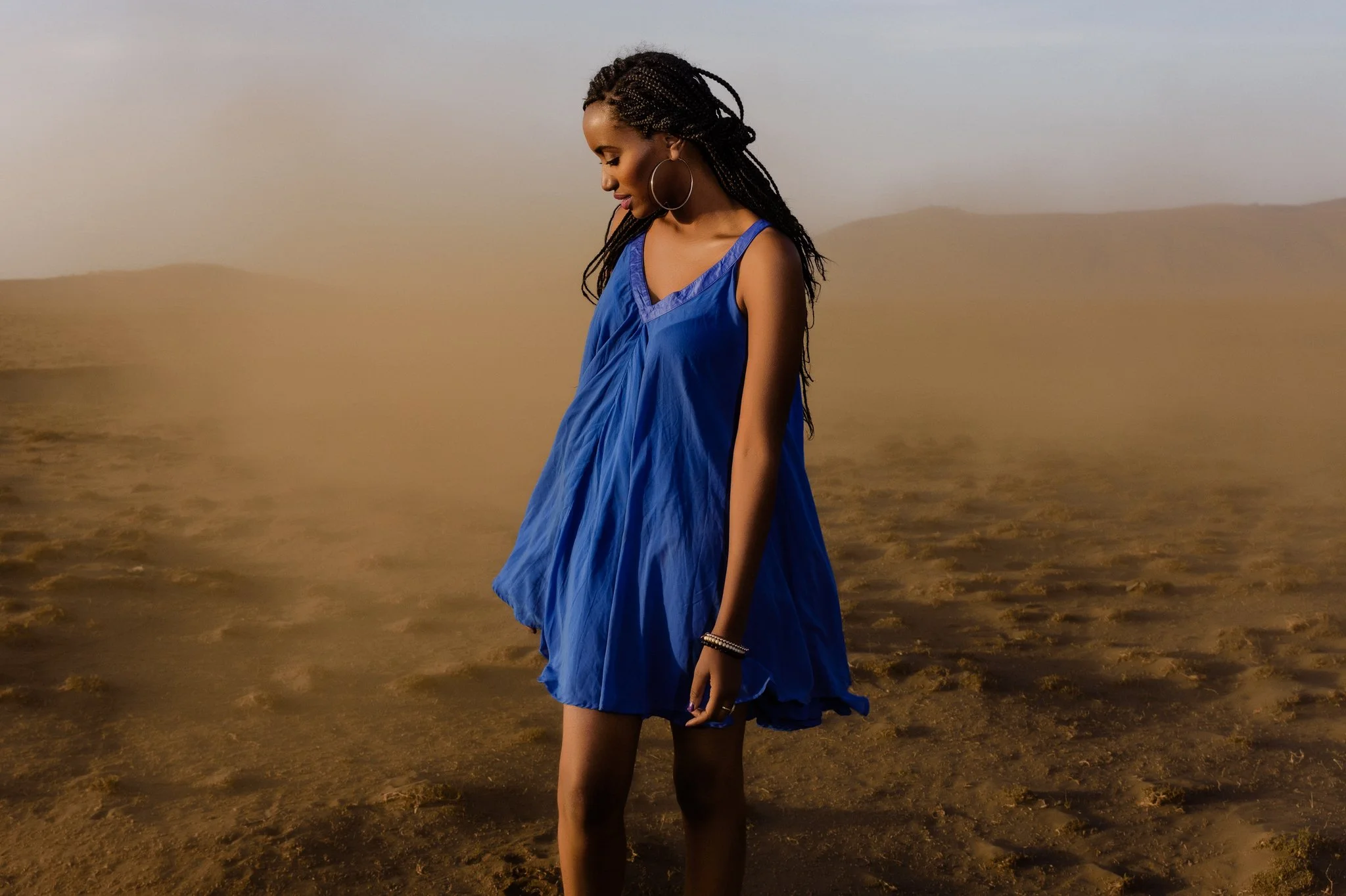 A woman with long braids and hoop earrings is standing in a desert landscape in Arusha, Tanzania, wearing a loose blue dress.