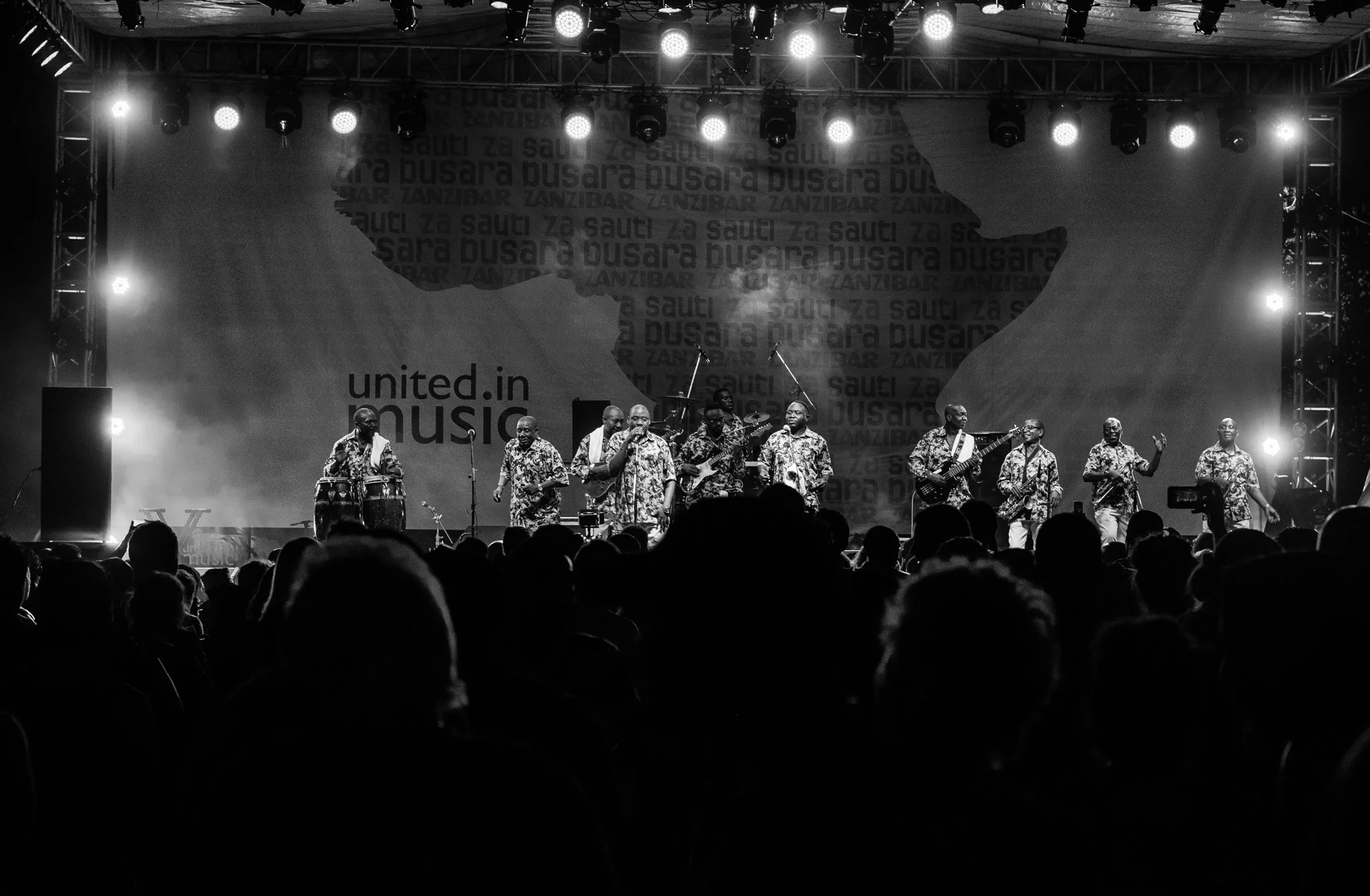 A musical performance on a stage with a band of ten members dressed in traditional patterned clothing, playing instruments including drums, guitars, and keyboard, with audience silhouettes in the foreground and a backdrop that features the text 'unit