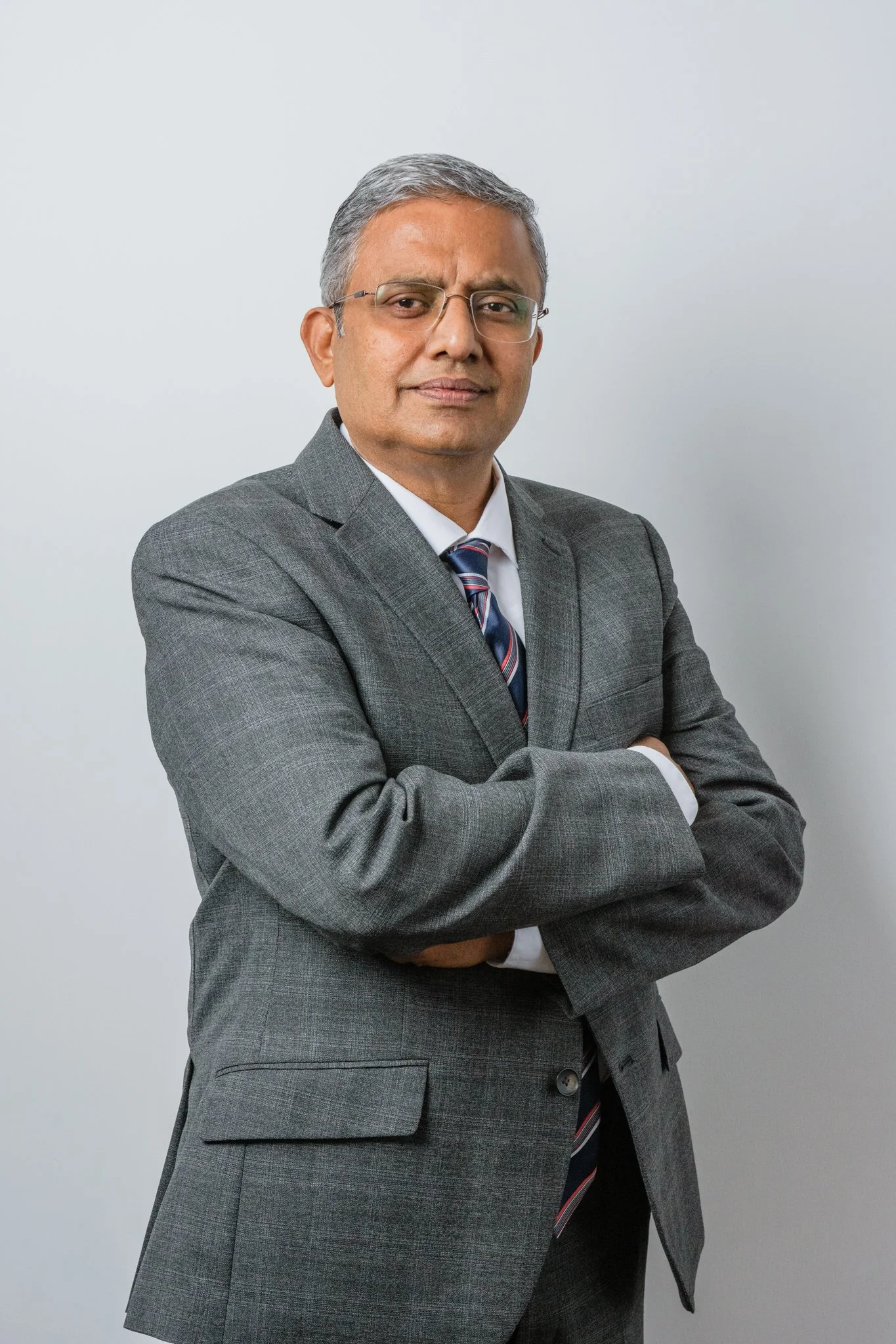 A professional man in a gray suit with glasses, sitting against a plain light background, arms crossed, looking at the camera.