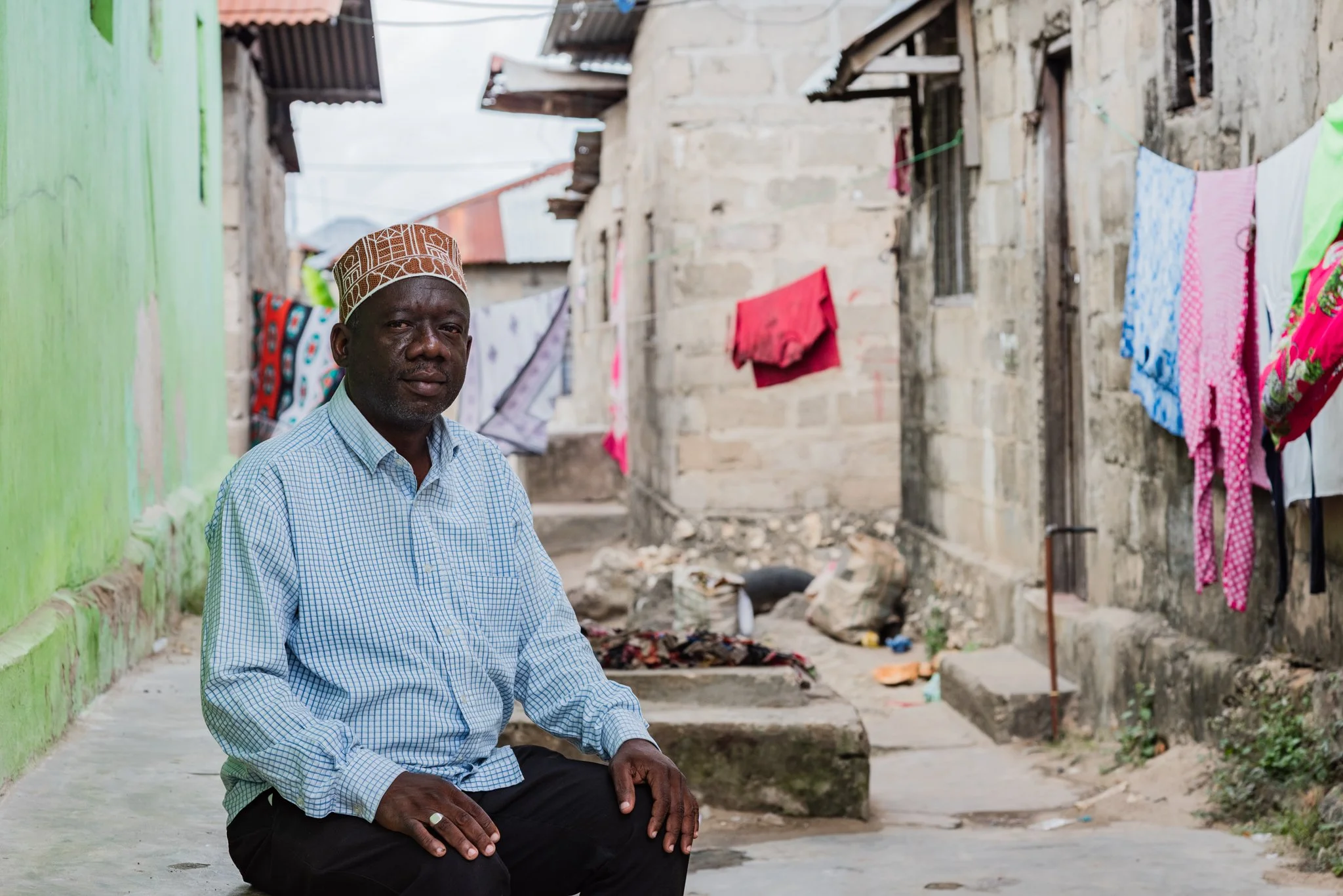 A man wearing a traditional cap and a checkered shirt, sitting on a concrete step in a residential area with clothes hanging to dry outside the houses in Zanzibar, Tanzania.
