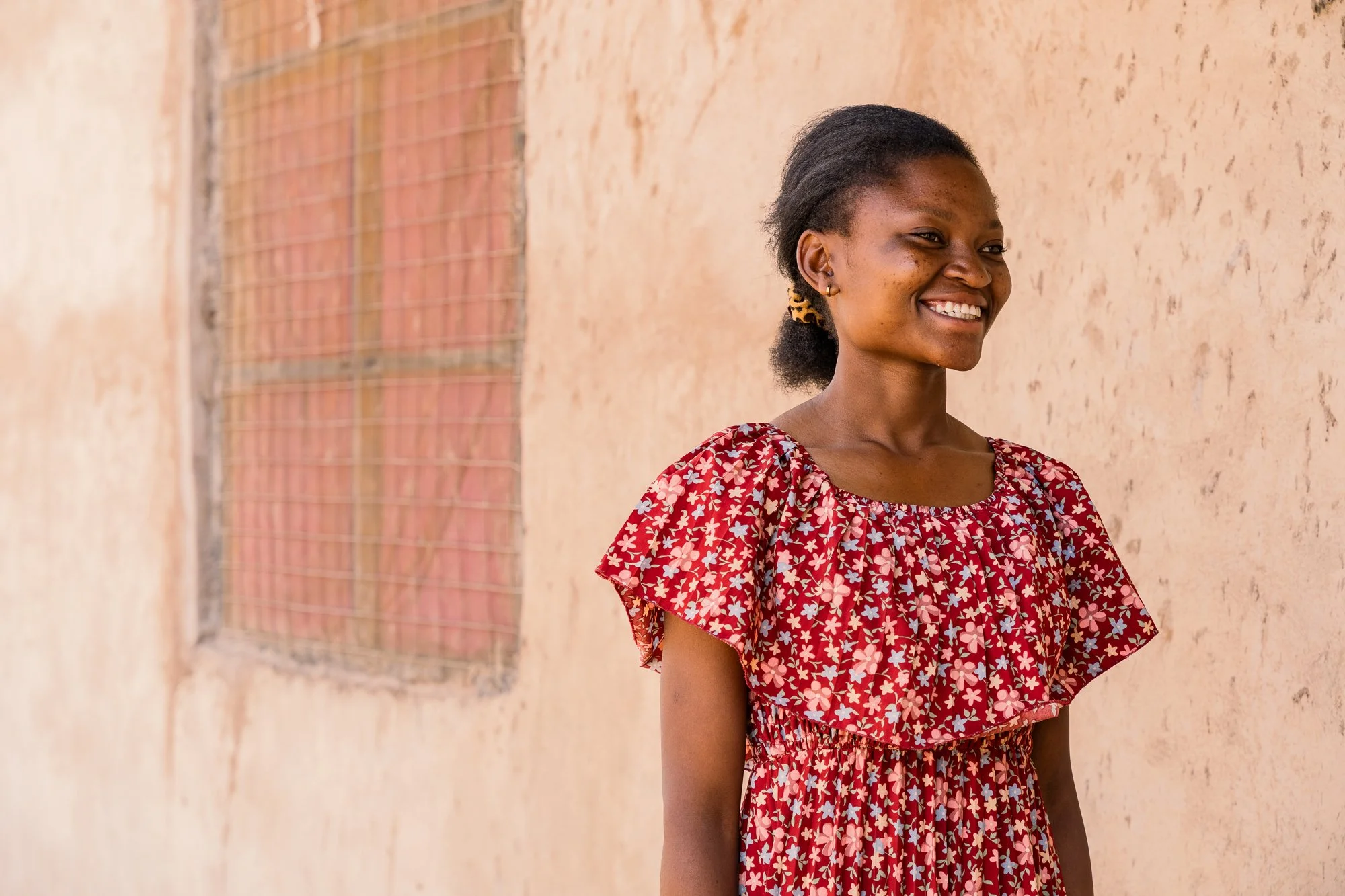 Documentary portrait of a young woman smiling, wearing a red floral dress and standing near a textured beige wall in Dodoma, Tanzania.