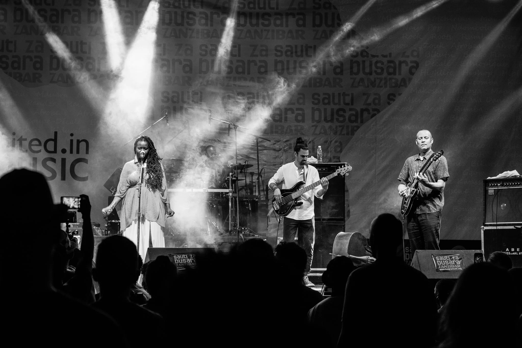 A black-and-white photo of a live music performance on stage with two guitarists, a bassist, and a female singer, surrounded by smoke and stage lights, with audience silhouettes in the foreground at the Sauti za Busara Music Festival in Zanzibar 2018