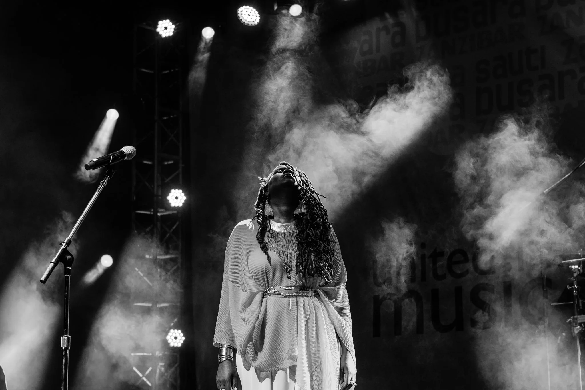 A woman with long dreadlocks singing on stage, illuminated by spotlights and surrounded by stage smoke, with a microphone stand nearby in Zanzibar at the Sauti za Busara Music Festival in 2018.