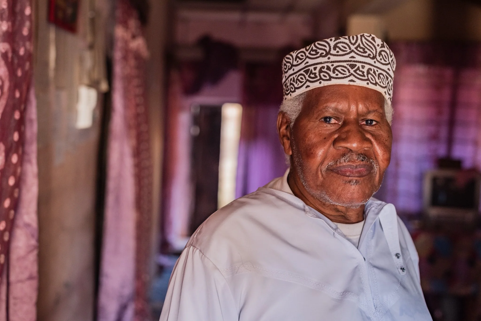 Portrait of an elderly man in a white traditional garment and a kufi, standing in a room with pink curtains and brick walls, looking at the camera in Zanzibar, Tanzania.