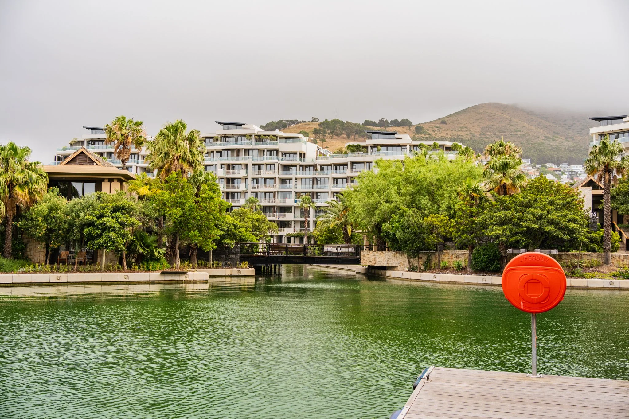 A waterfront scene with a dock, lush green trees, and a modern apartment complex in the background, with mountains partially obscured by clouds.