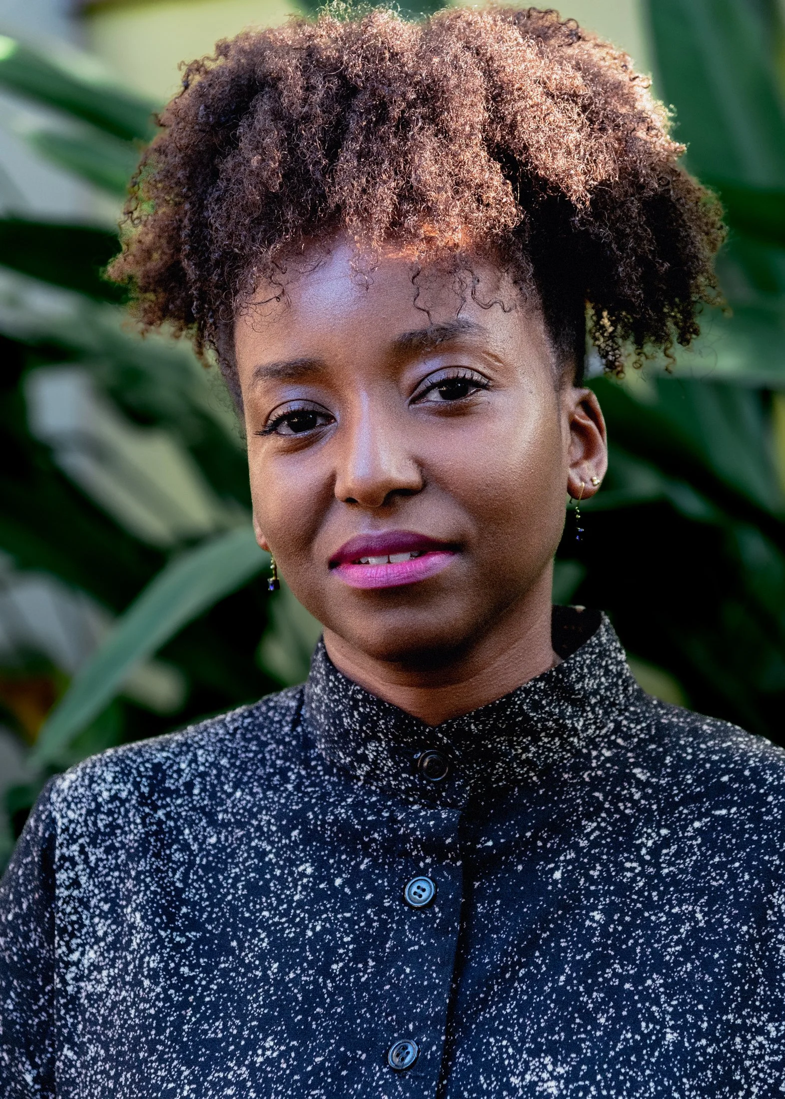 Close-up portrait of a woman with natural curly hair, wearing a dark, patterned button-up shirt, standing outdoors with large green leaves in the background.