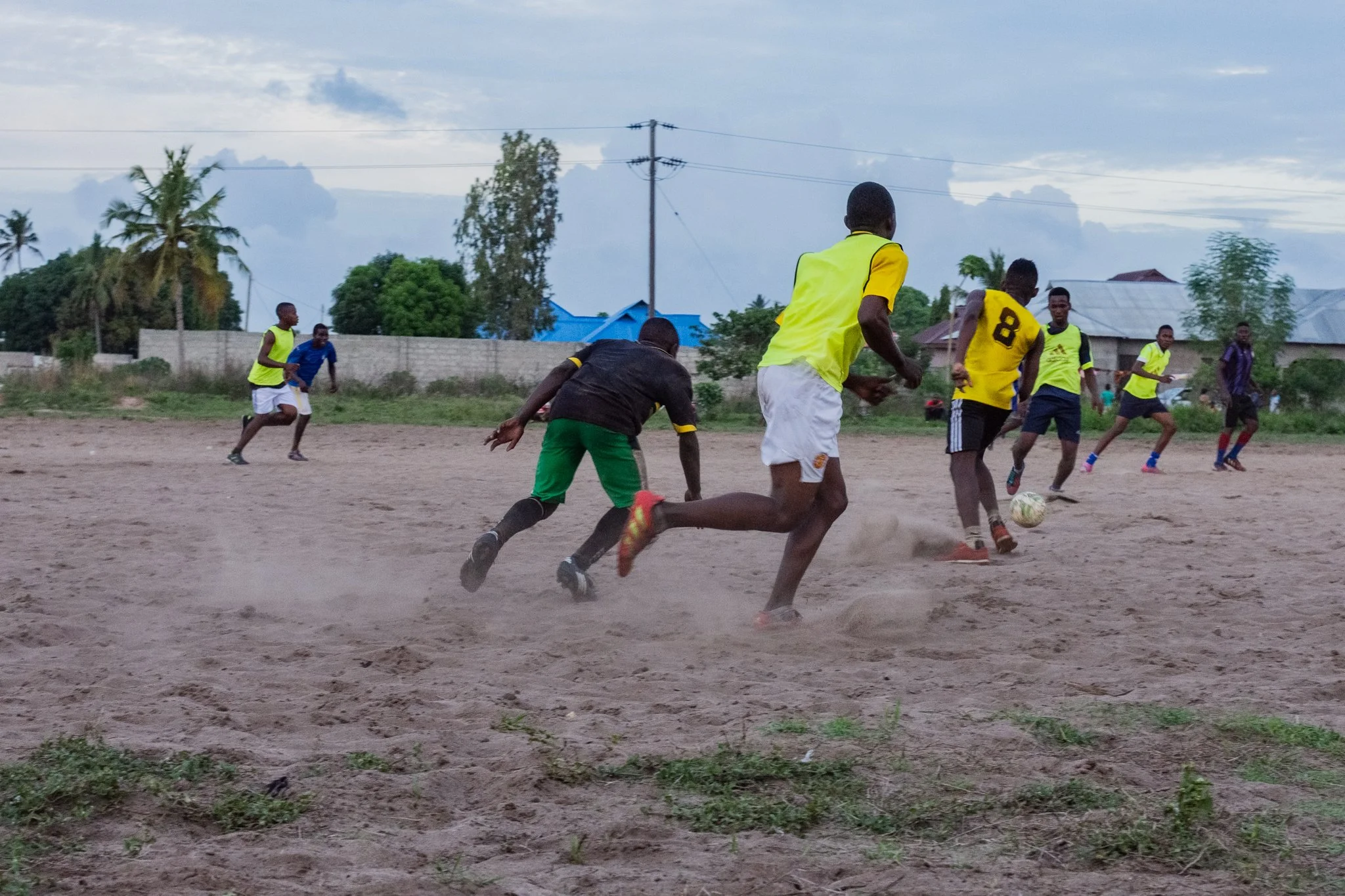 A group of young men playing football on a sandy field in a residential area, some wearing yellow jerseys and others in darker clothing, with trees and houses in the background in Toangoma, Dar es Salaam.