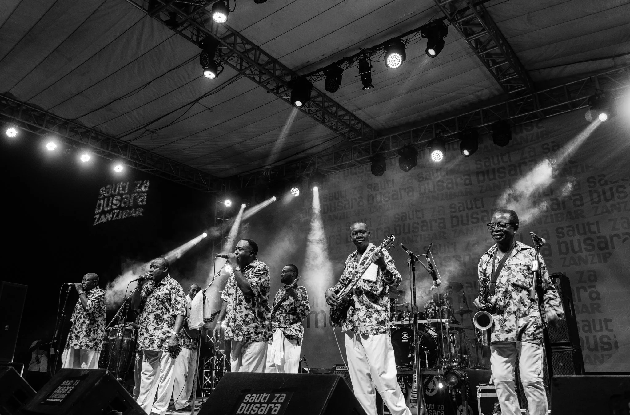 A black and white photo of a musical band performing on stage, with seven members wearing patterned shirts, singing, playing instruments, and smiling in Zanzibar at the Sauti za Busara Music Festival in 2018.