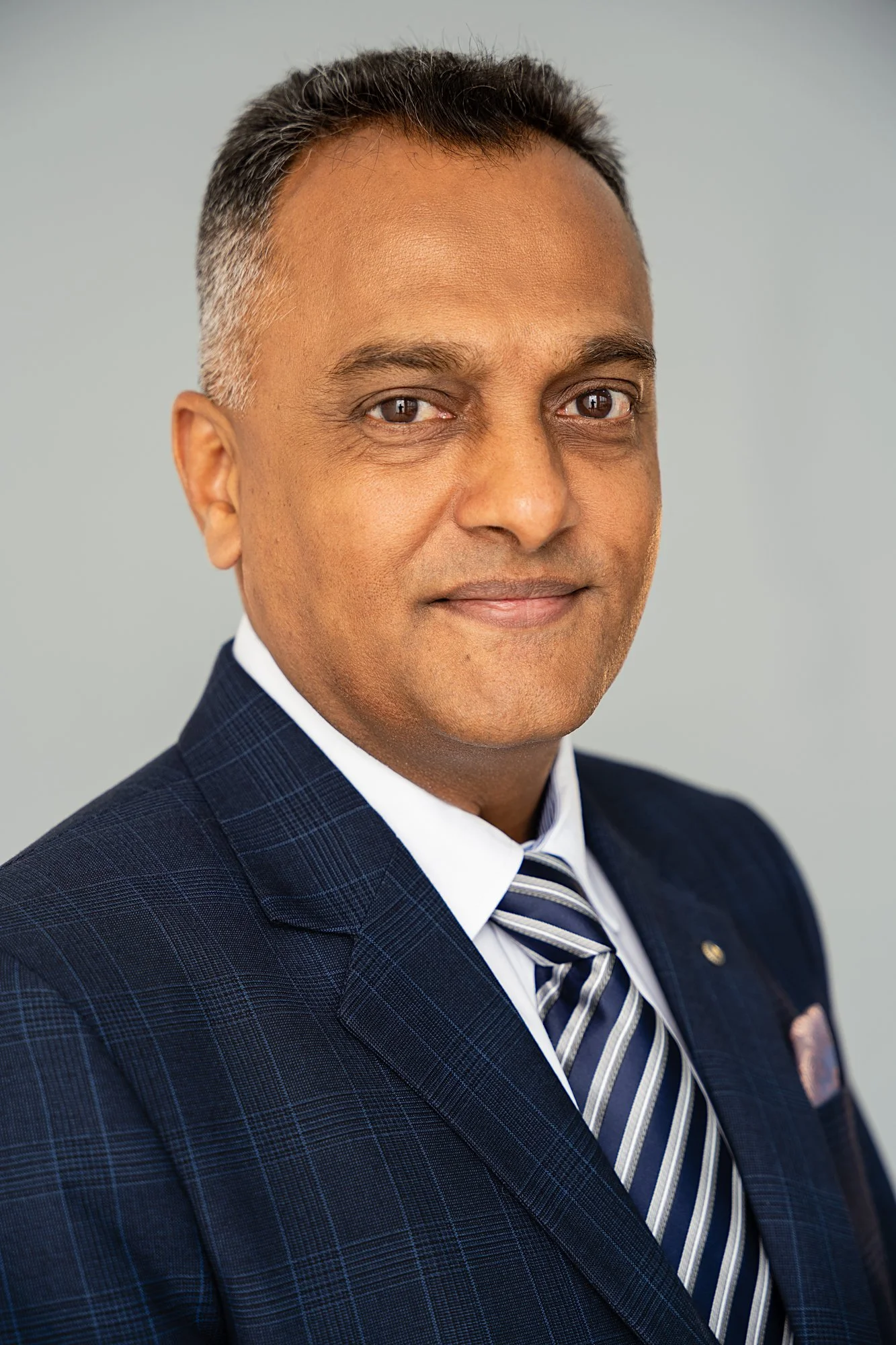 A professional man in a dark blue checkered suit with a white shirt and a blue striped tie posing against a plain light gray background.