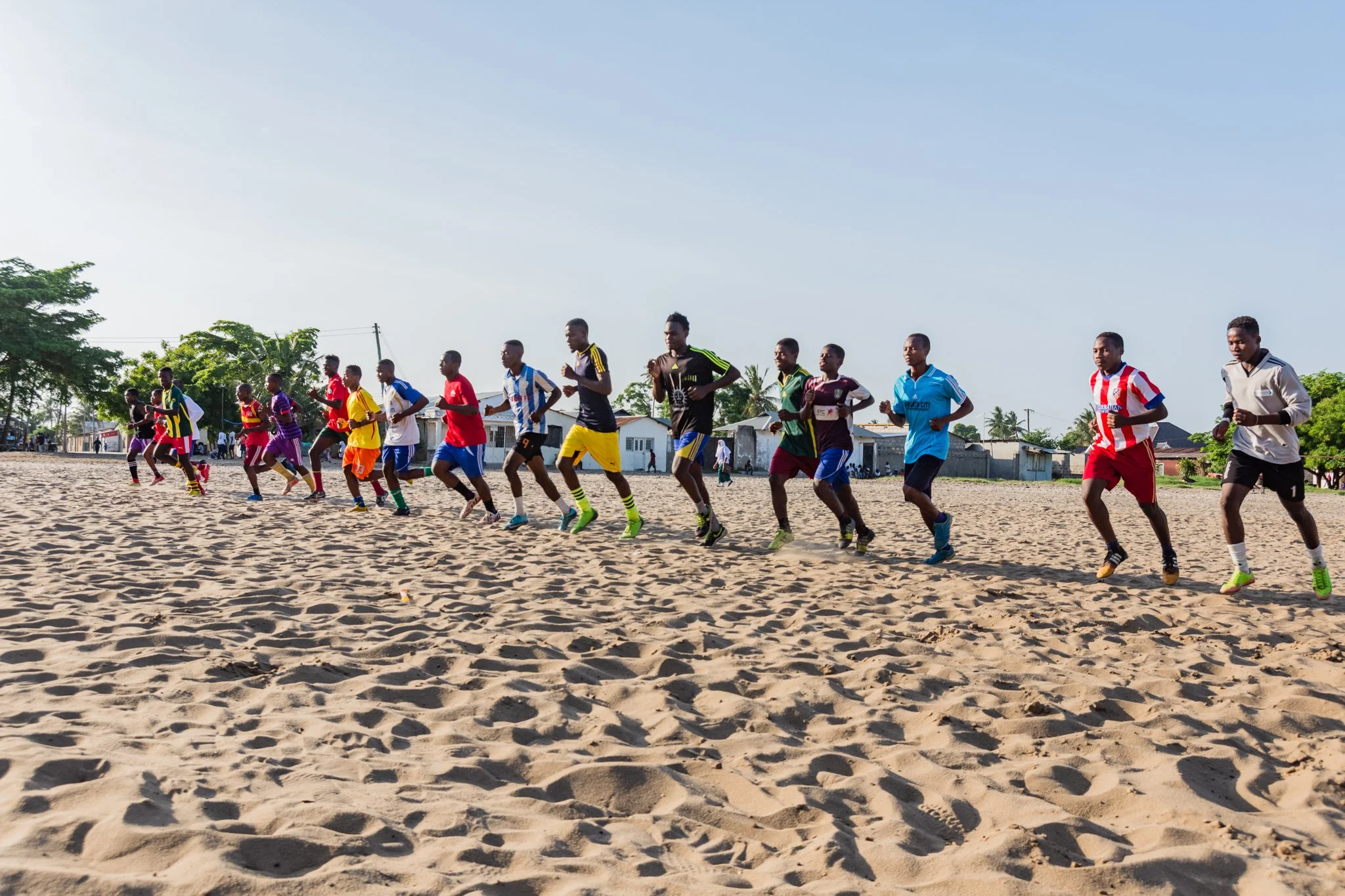 A group of young men running on a sandy field, some wearing colourful sports jerseys and shorts, with houses and trees in the background under a clear sky in Mbagala, Dar es Salaam.