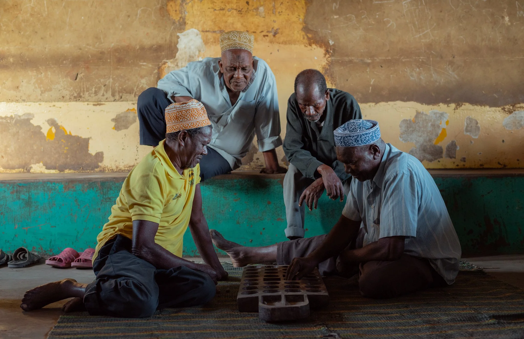 Four elderly men play a traditional game of bao, sitting and kneeling on a woven rug against a weathered yellow wall in Zanzibar, Tanzania.