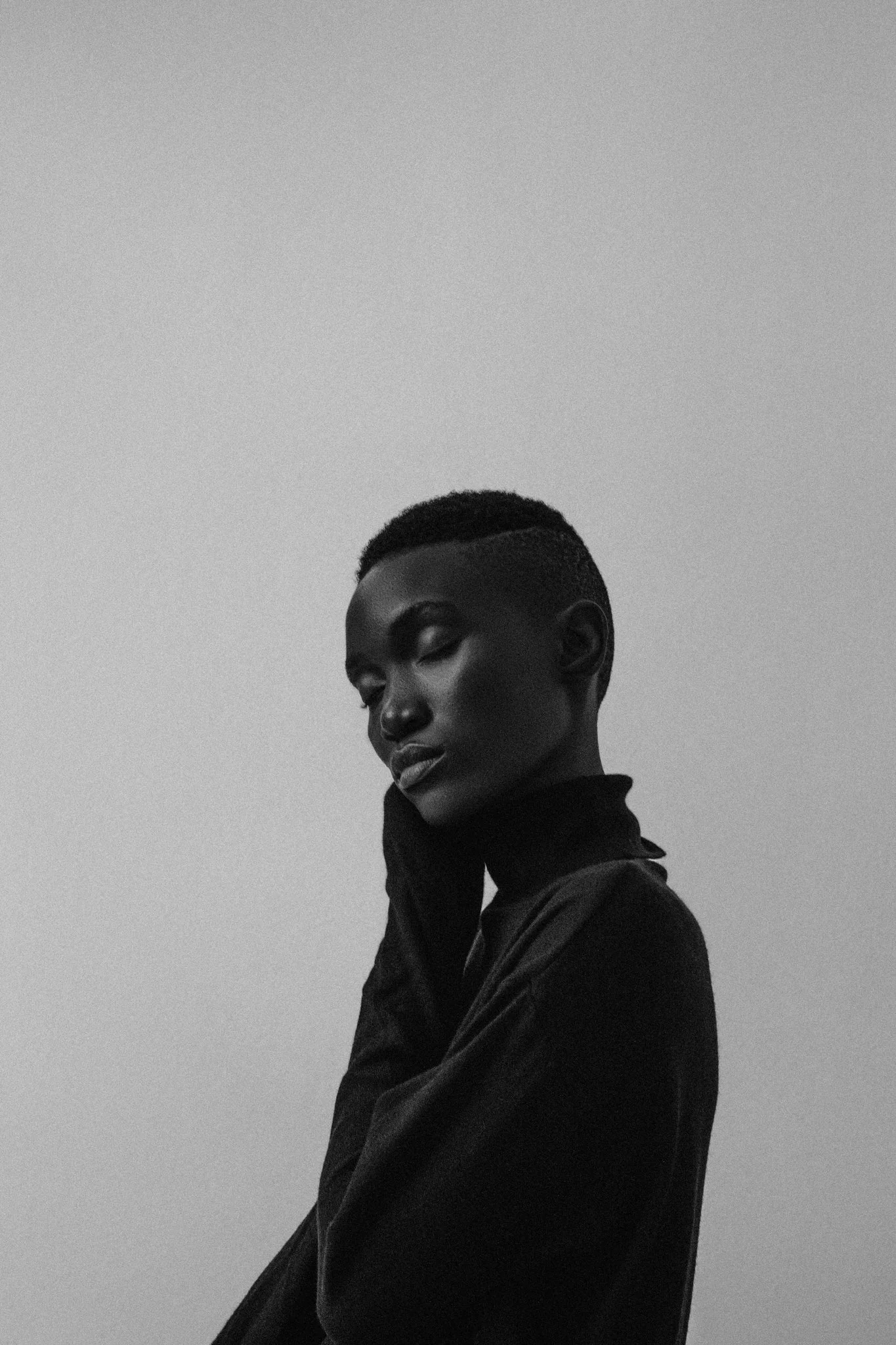 A high-contrast, black and white fine art portrait of a black female model with short hair, wearing a high-neck top, with her hand resting on her face, looking thoughtful.