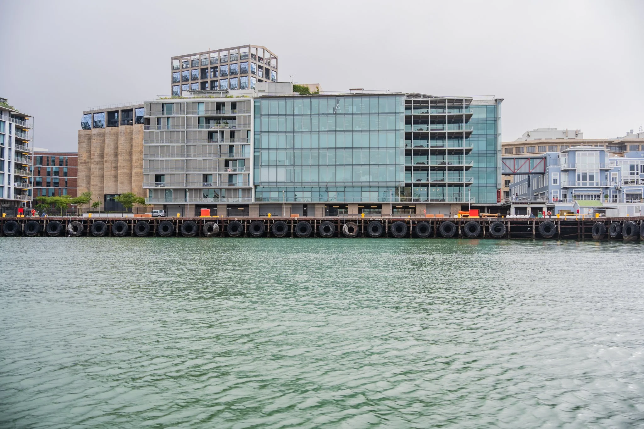 Modern waterfront buildings along a pier with calm water in the foreground.