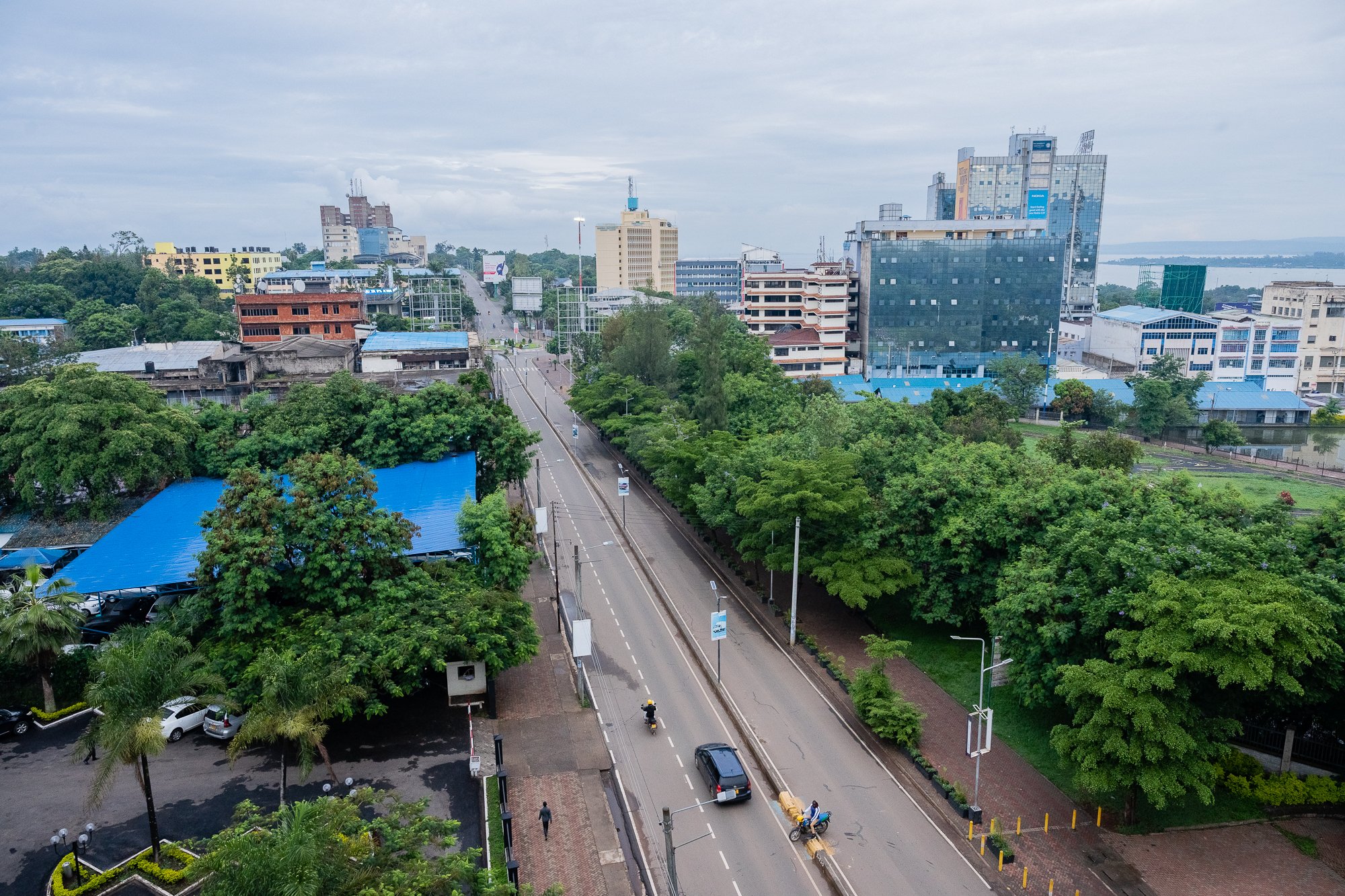 Overhead view of a city street with cars and motorbikes, surrounded by lush green trees and tall modern buildings under a cloudy sky. Kisumu, Kenya.