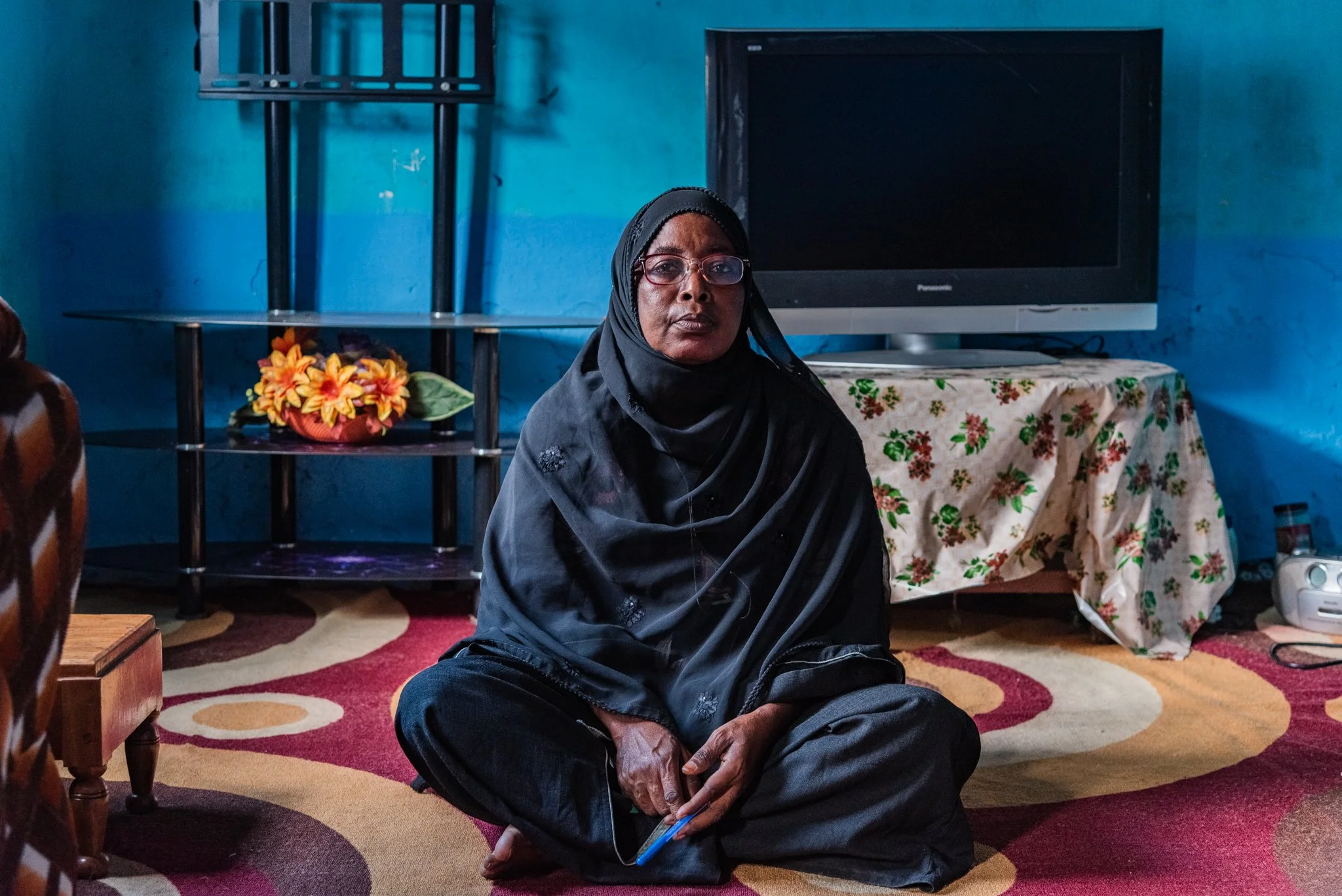 An older woman in a hijab on the floor of a room in Zanzibar, Tanzania, with a red-and-beige patterned carpet, a blue wall, a TV on a stand covered with a floral cloth, and a small flower arrangement on a glass shelf behind her.