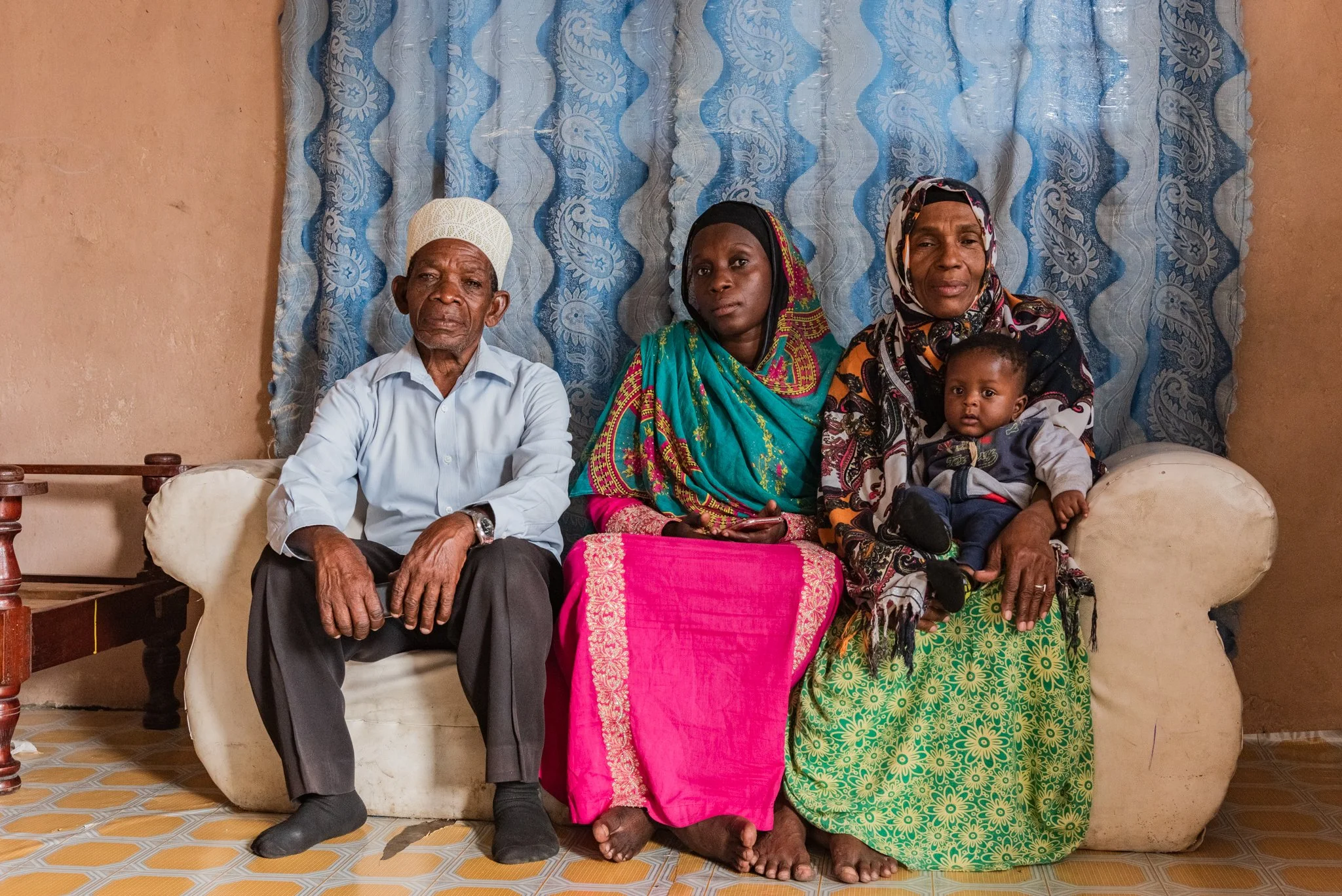 A family of four sitting on a sofa, including an elderly man, two women, and a baby, with a blue-patterned curtain backdrop in Zanzibar, Tanzania.