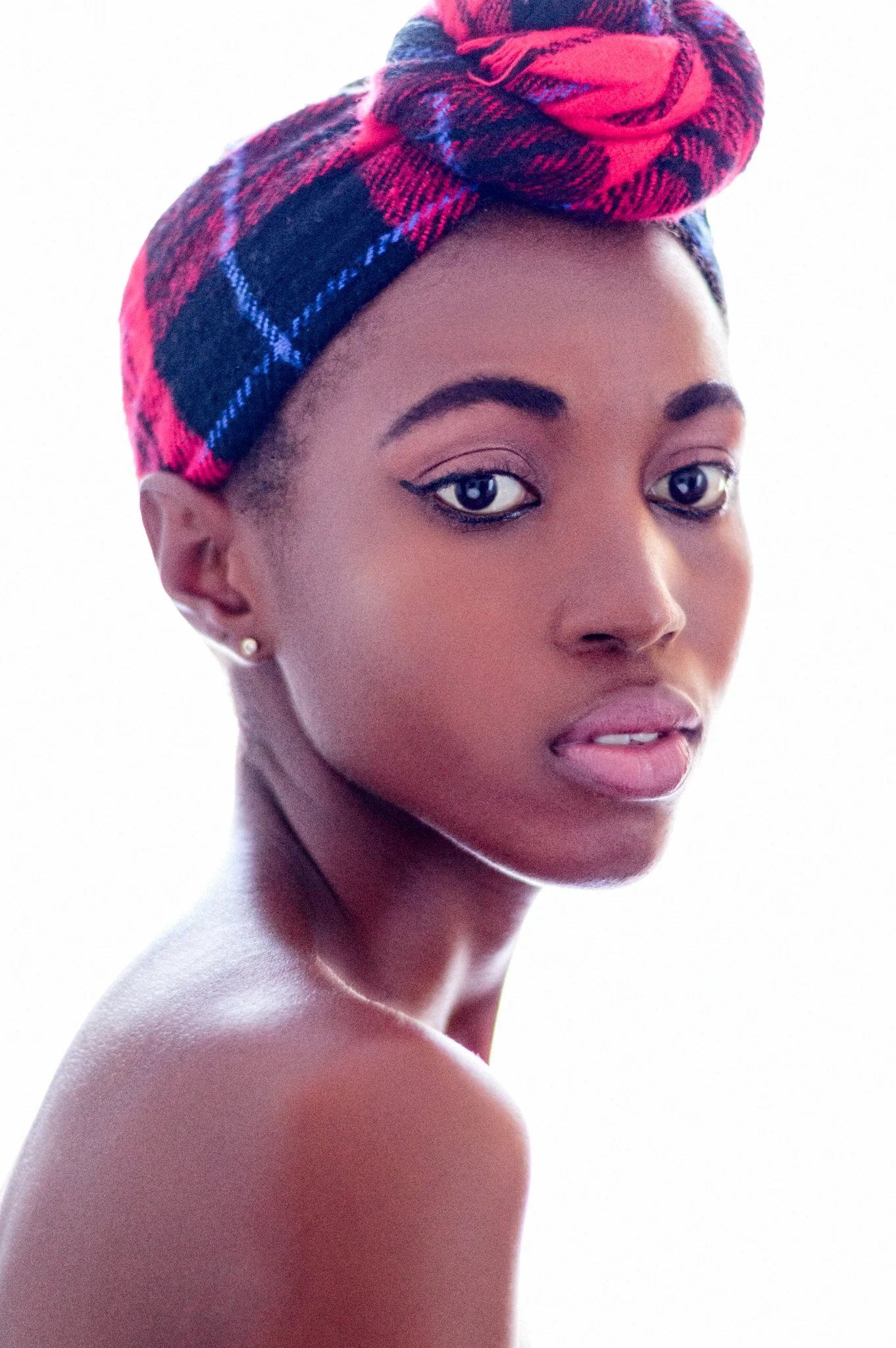 Portrait of a young woman with dark skin, short hair, wearing a colorful pink and black plaid head wrap, looking over her shoulder against a white background.