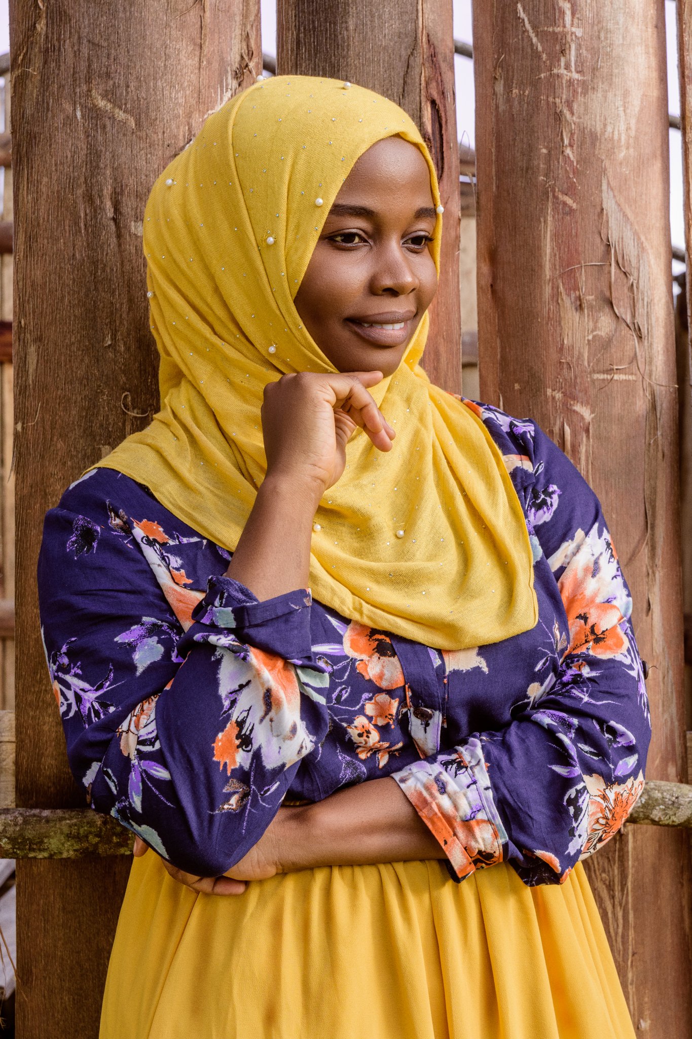 A woman wearing a yellow hijab with pearl embellishments and a floral navy blue top with orange, white, and purple flowers, standing against a wooden fence.