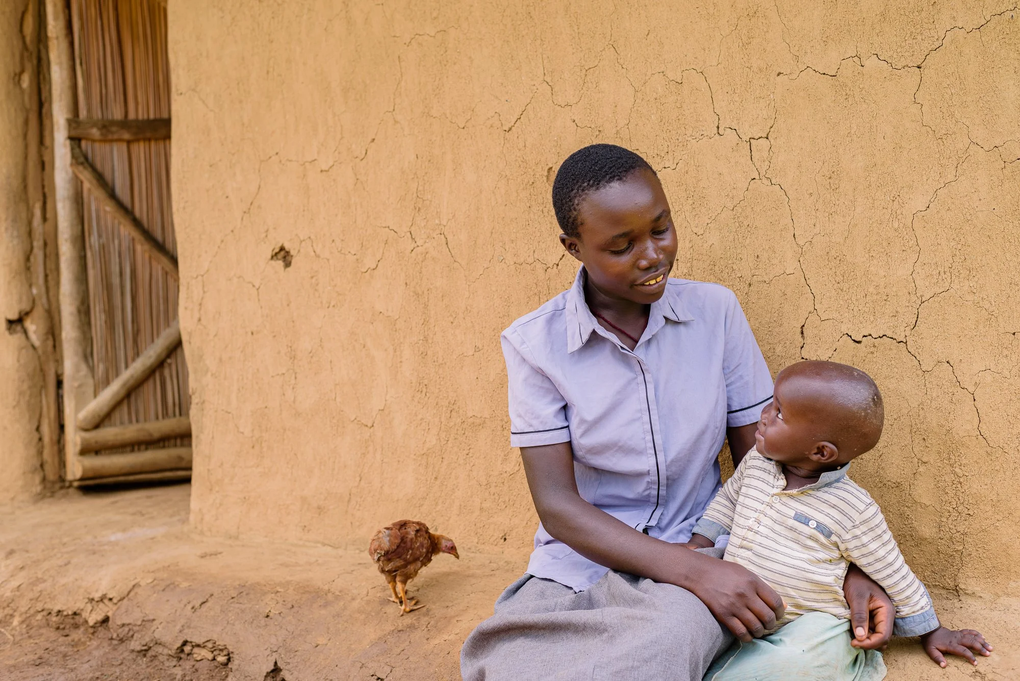 Girl and child sit outside a clay wall, with a chicken on the ground nearby in Siaya County, Kenya.