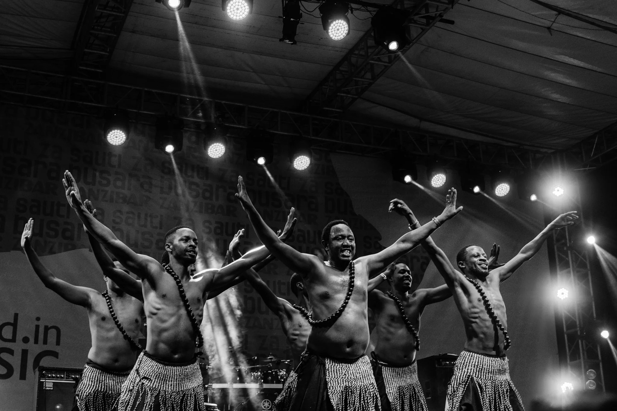 A group of six shirtless male performers in traditional attire dancing on stage with arms extended and smiling, under stage lights in Zanzibar at the Sauti za Busara Music Festival in 2018.