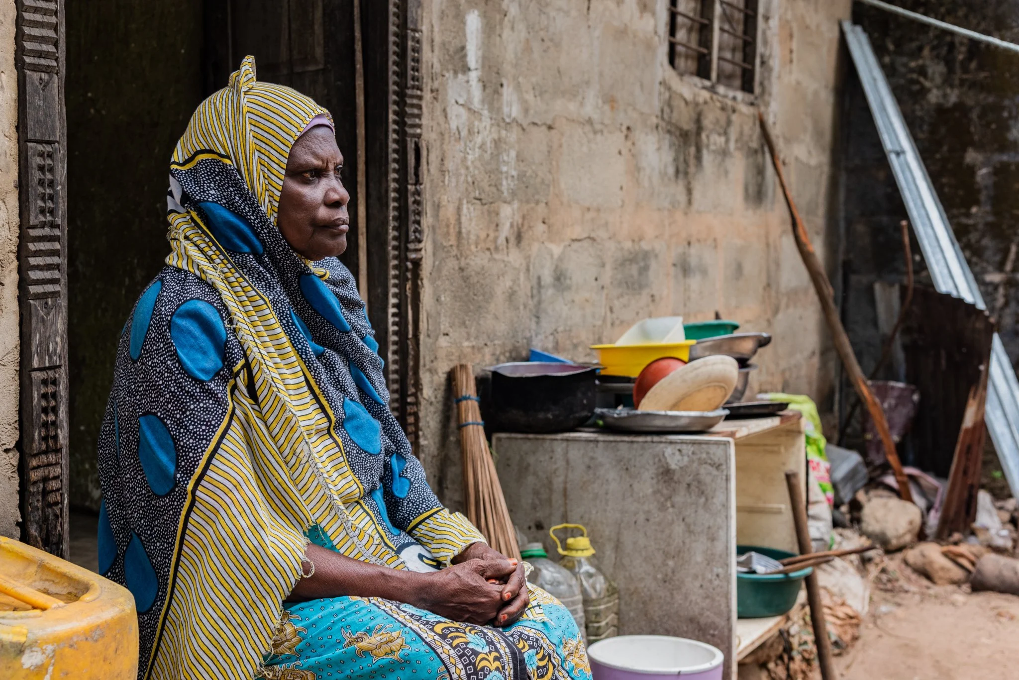 An elderly woman sitting outside near a worn, muddy wall with scattered dishes and kitchen items in Zanzibar, Tanzania.