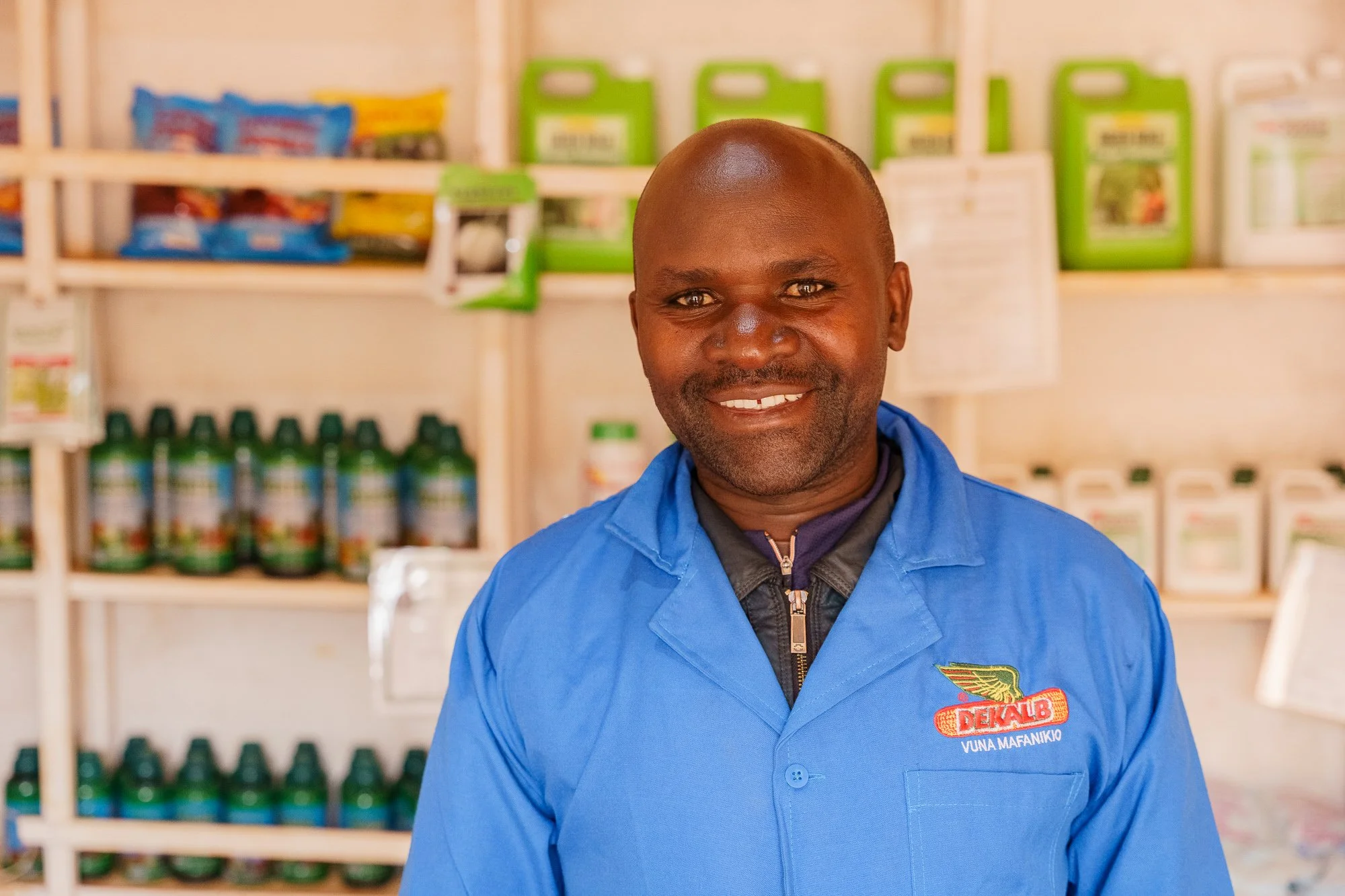 A man in a blue DeKalb-branded uniform standing in front of stocked shelves inside an agro-input shop during a Farm Africa assignment in Babati, Manyara, Tanzania.