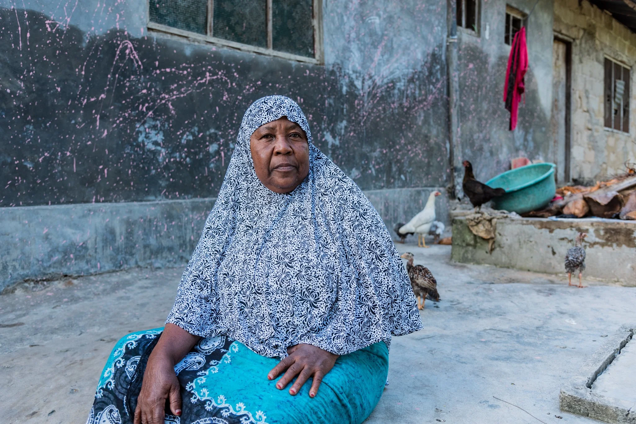 An older woman sitting outside in front of a weathered building with chickens around her in Zanzibar, Tanzania.