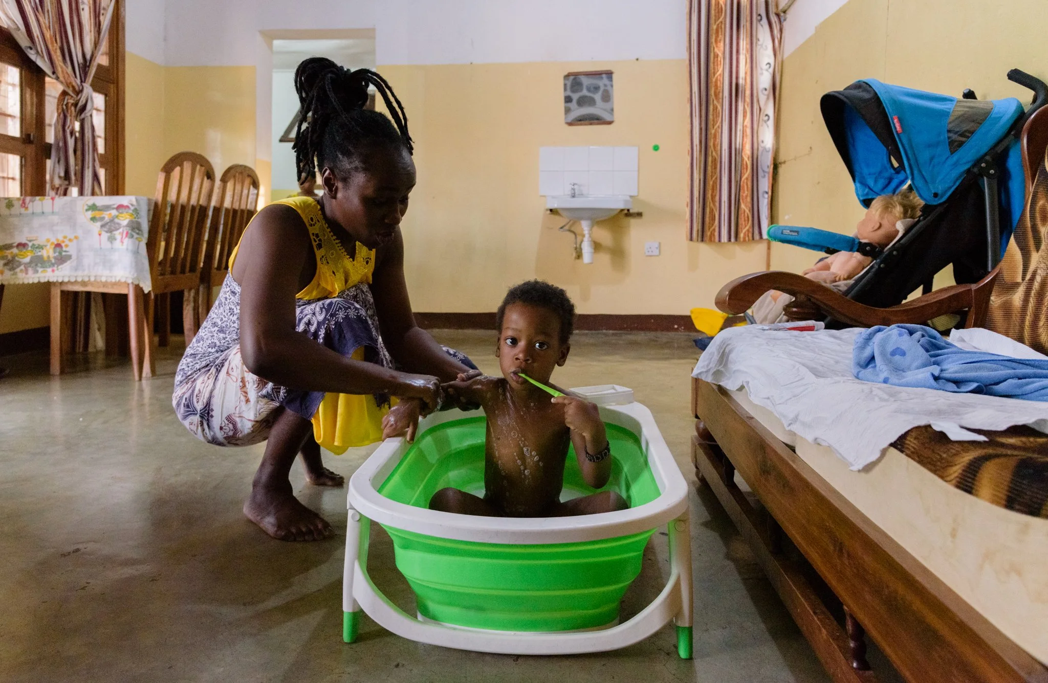 Woman bathing a young child in a small plastic tub inside a room in Mwanza, Tanzania, as the child holds a toothbrush and looks toward the camera.