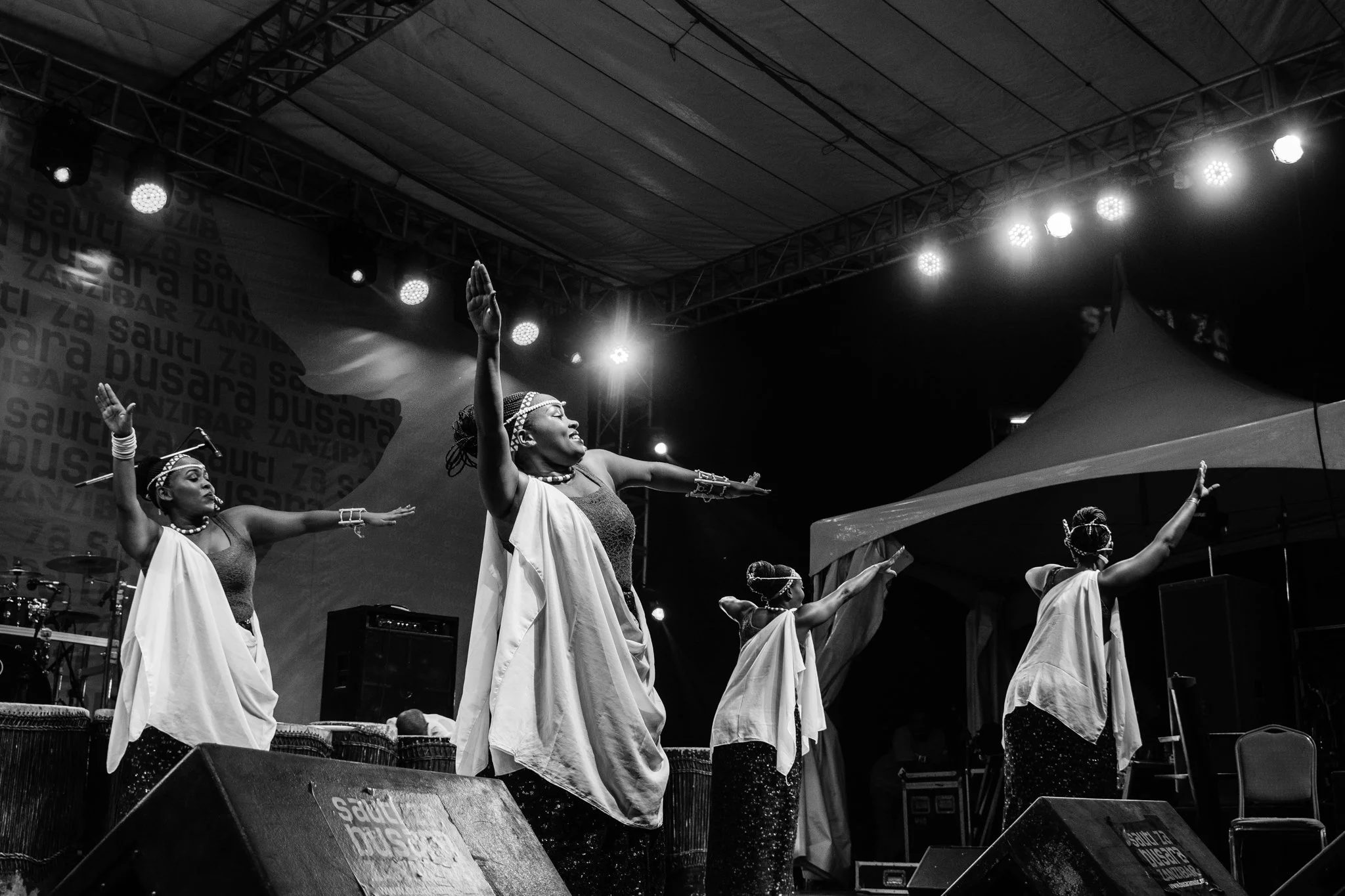Four women wearing traditional African attire and jewellery are performing a dance on stage, with arms raised and smiling, under stage lights at an outdoor event in Zanzibar at the Sauti za Busara Music Festival in 2018.
