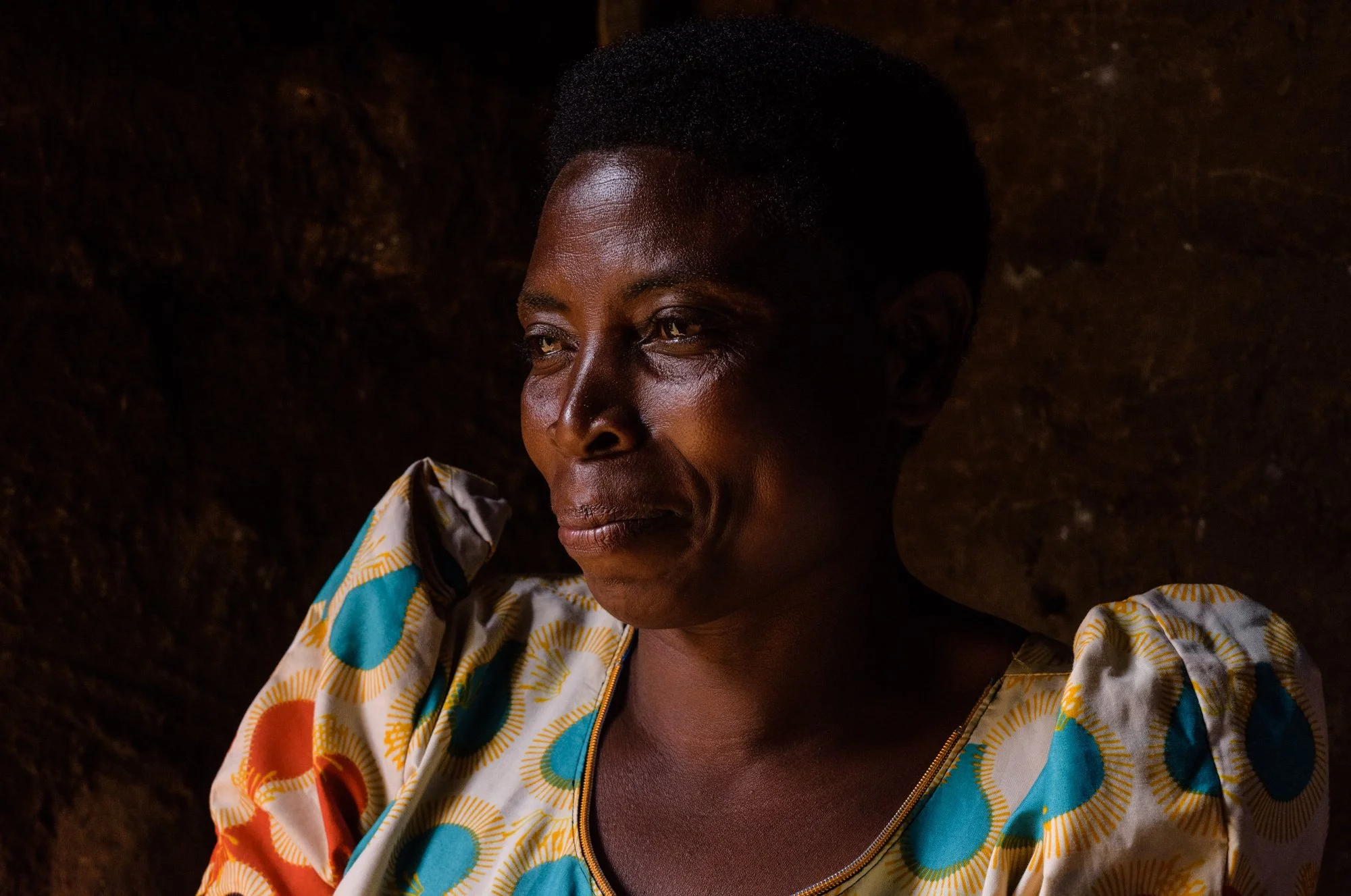 Documentary portrait of an elderly woman seated indoors, illuminated by soft natural light in Mbeya, Tanzania.