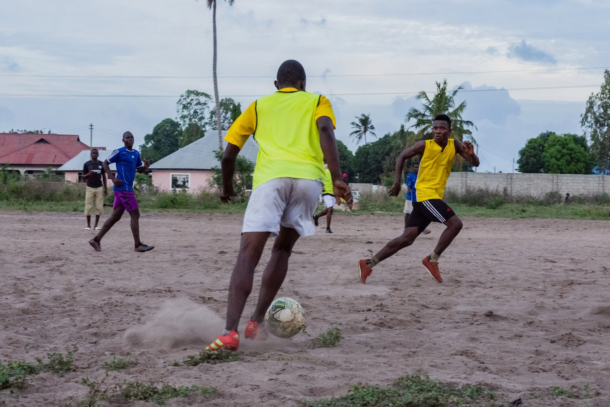 Young men playing football on a sandy field in a residential area, with houses and trees in the background in Toangoma, Dar es Salaam.