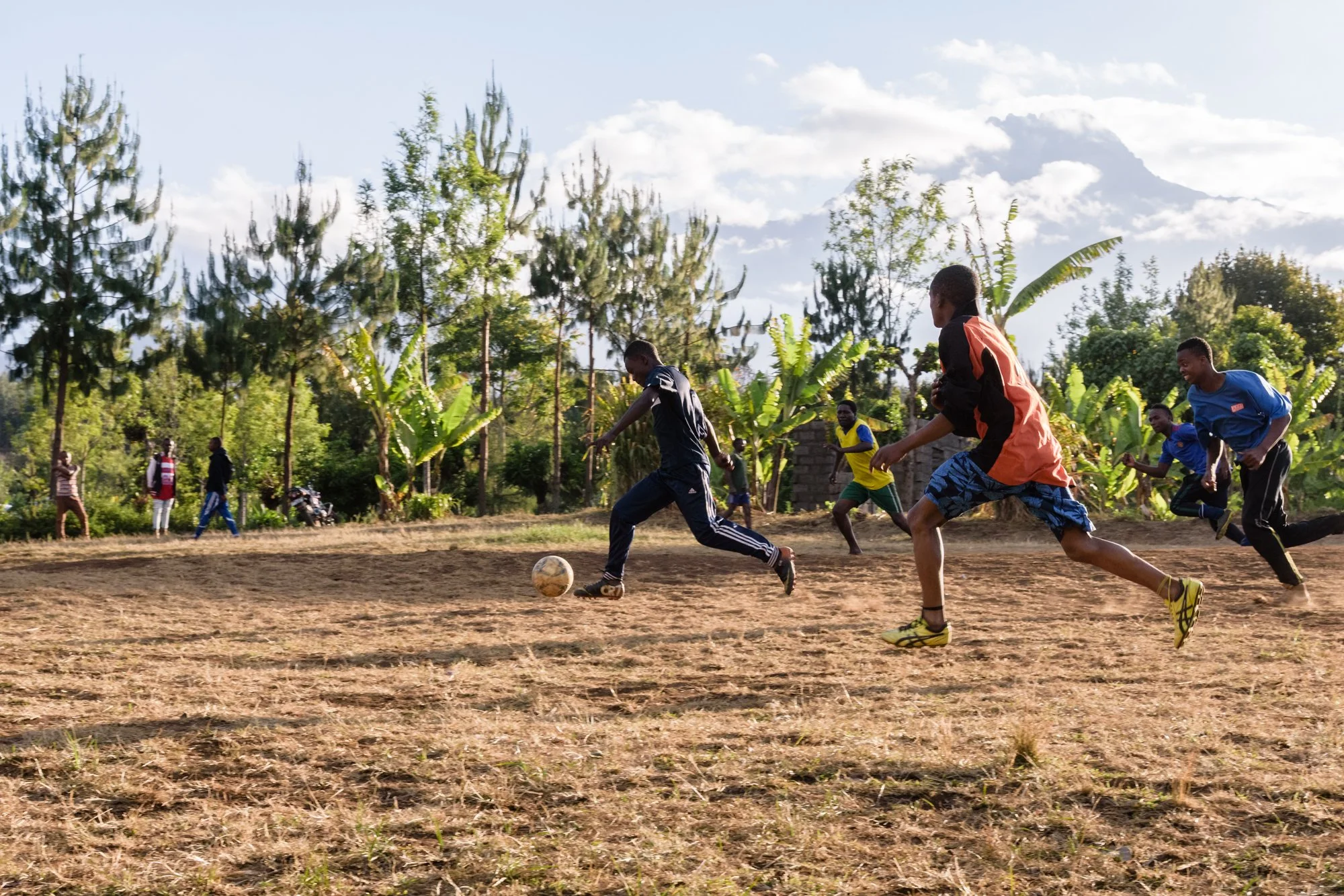 Young boys playing football on a field surrounded by trees, with spectators watching under a partly cloudy sky in Kilimanjaro Region, Tanzania.