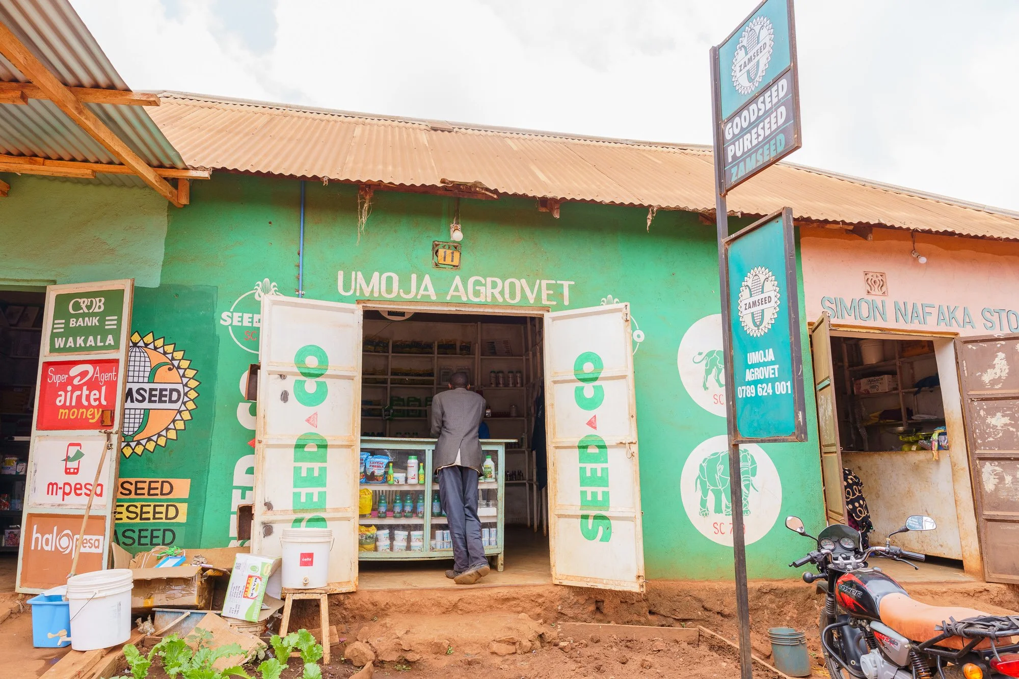 Exterior view of a small agro-input store with branded signboards, open doors, and agricultural products on display, with a motorcycle parked outside in Babati, Manyara, Tanzania.