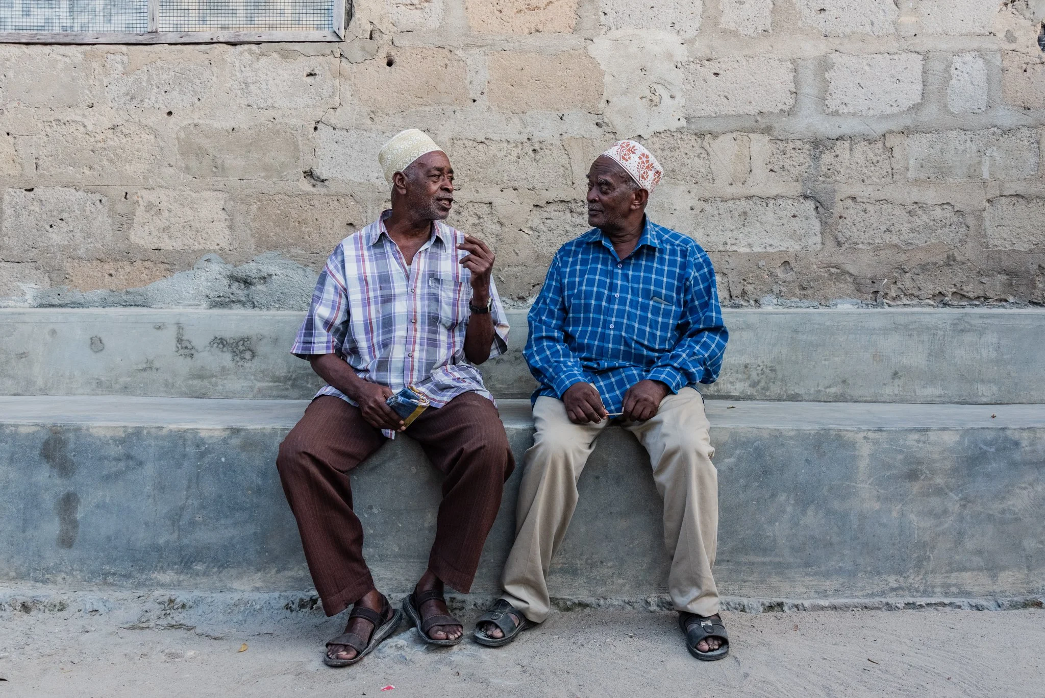 Two elderly men sitting on a concrete bench against a brick wall, engaged in conversation. Both wearing patterned shirts, light-colored trousers, sandals, and a kufi in Zanzibar, Tanzania.