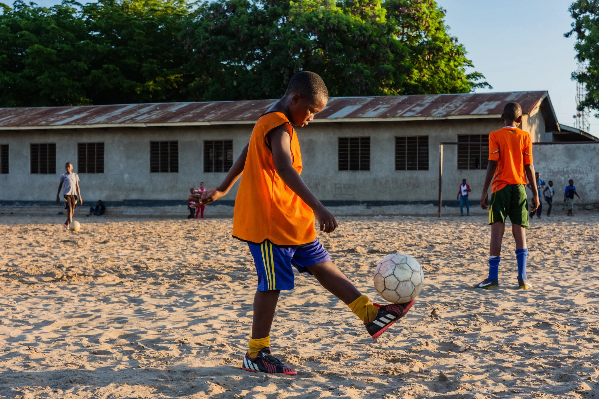 Children playing soccer on a dirt field with a school building and trees in the background during daylight in Mbagala, Dar es Salaam.