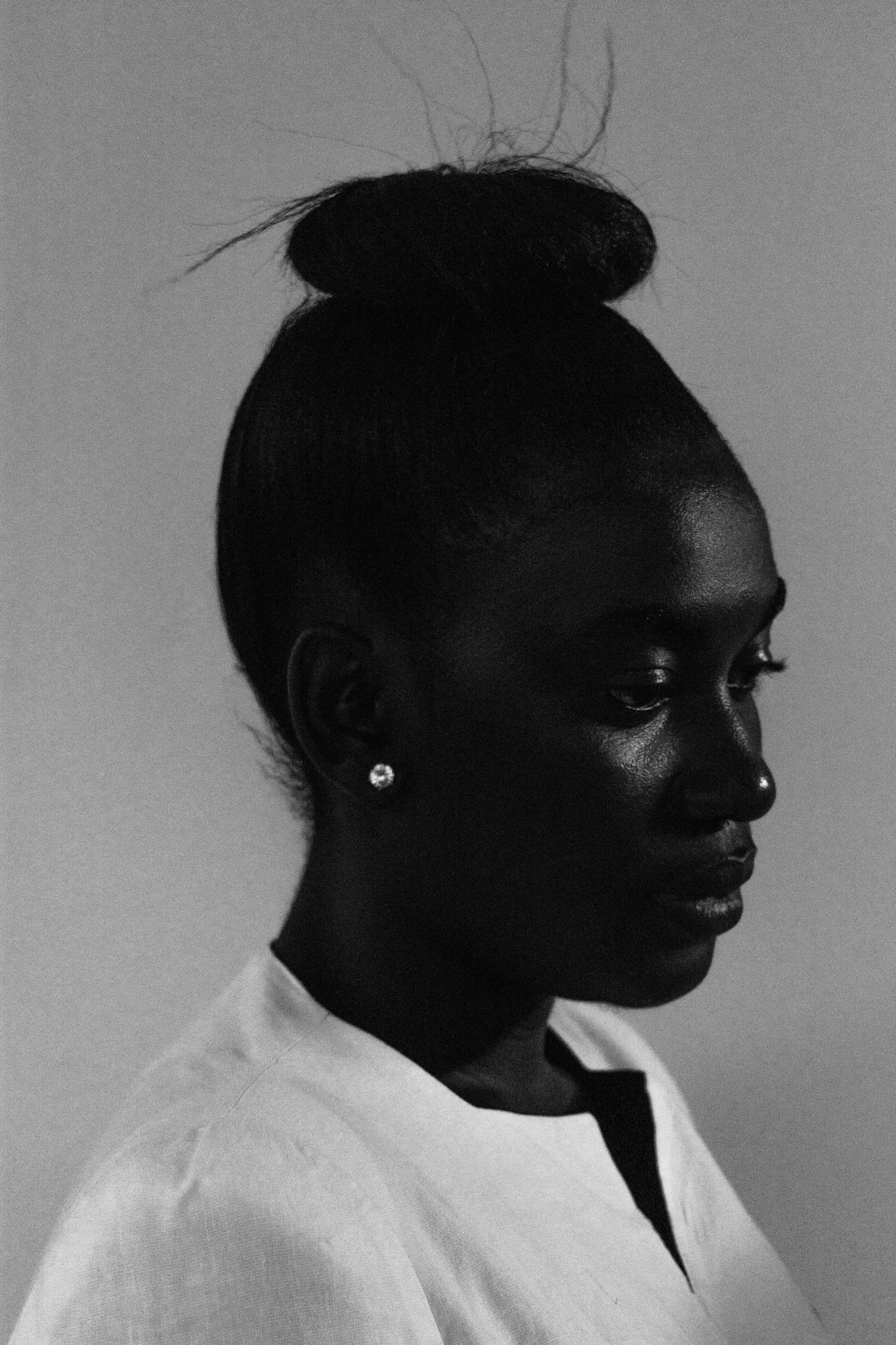 A high-contrast, black and white fine art side profile of a woman with her hair in a high bun, wearing earrings and a light-colored top.