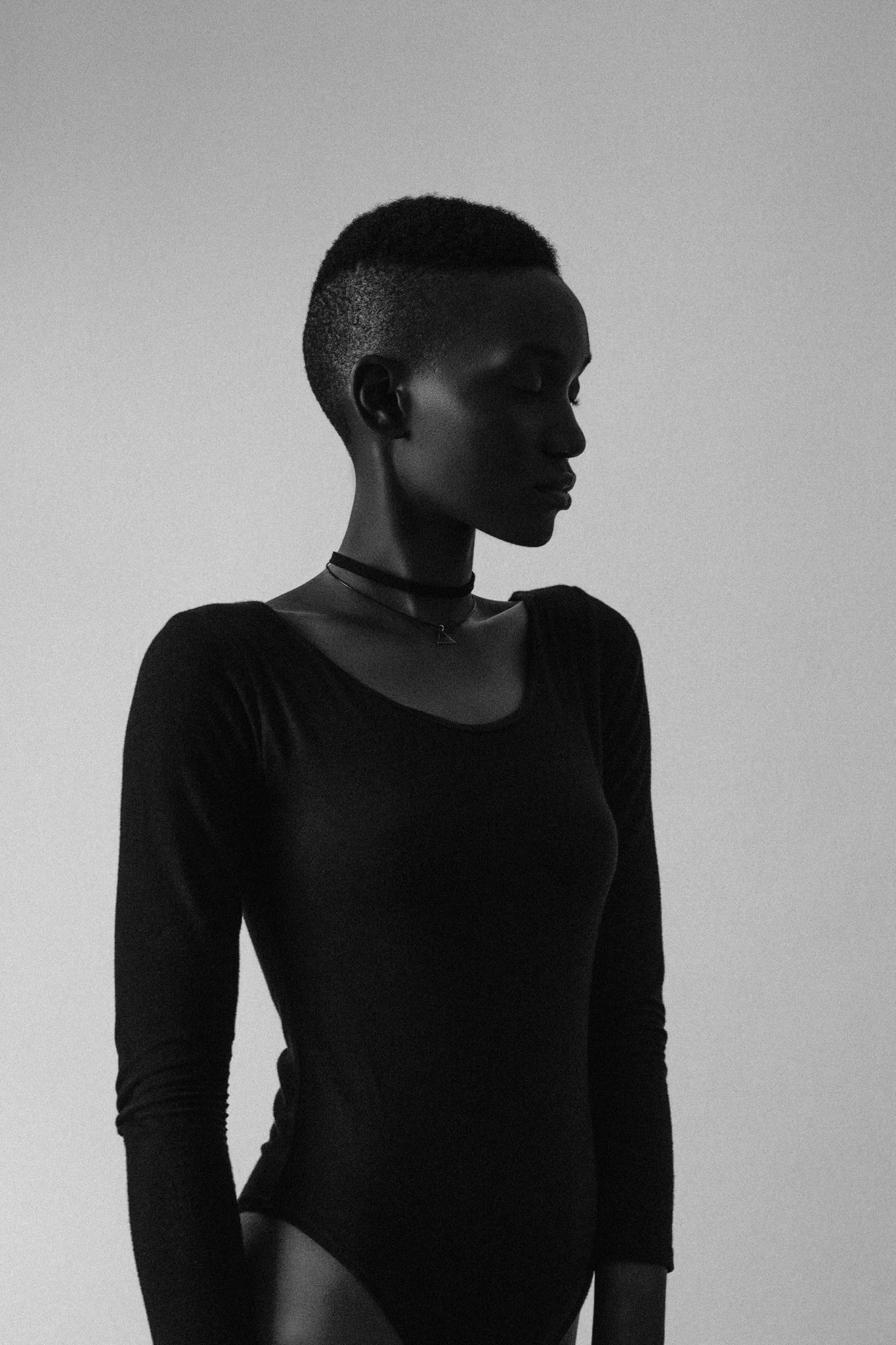 A high-contrast, black and white fine art photo of a female black model with a short haircut, wearing a long-sleeve black top, and standing against a plain background in Dar es Salaam, Tanzania.