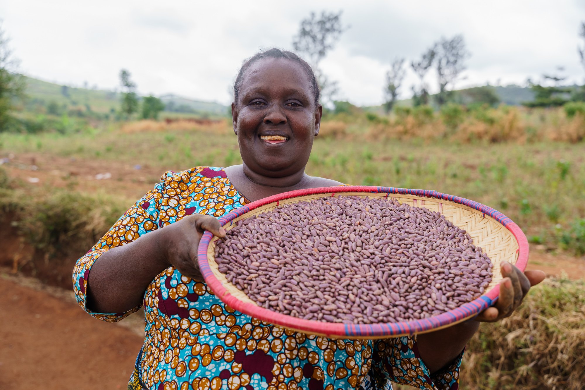 A smiling woman outdoors holding a large basket of purple beans in Arusha, Tanzania.