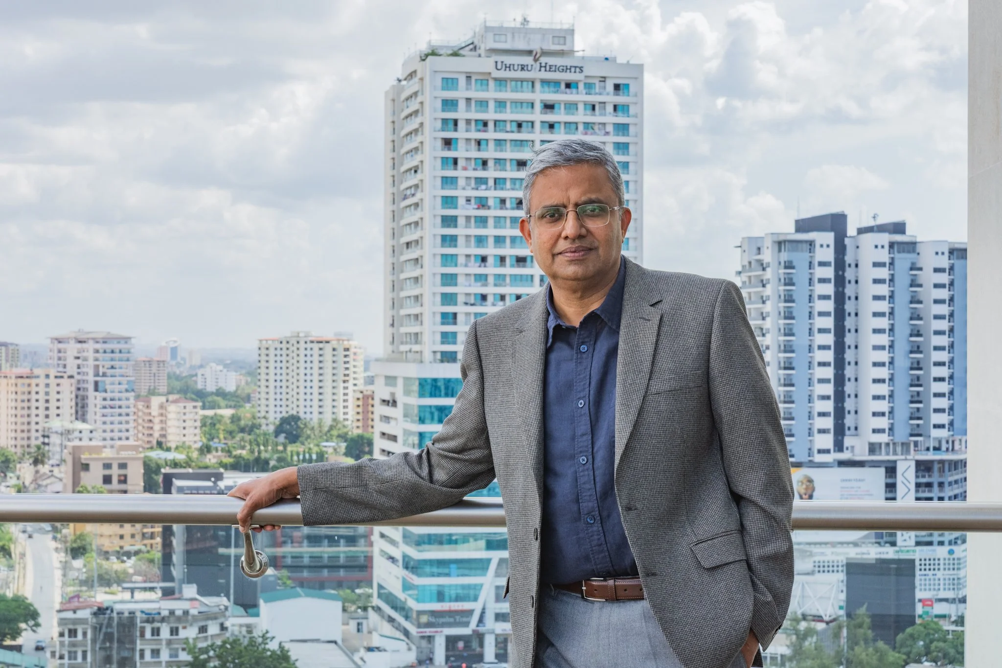A man in a gray business suit and glasses standing on a balcony with a cityscape of tall buildings behind him in Dar es Salaam.