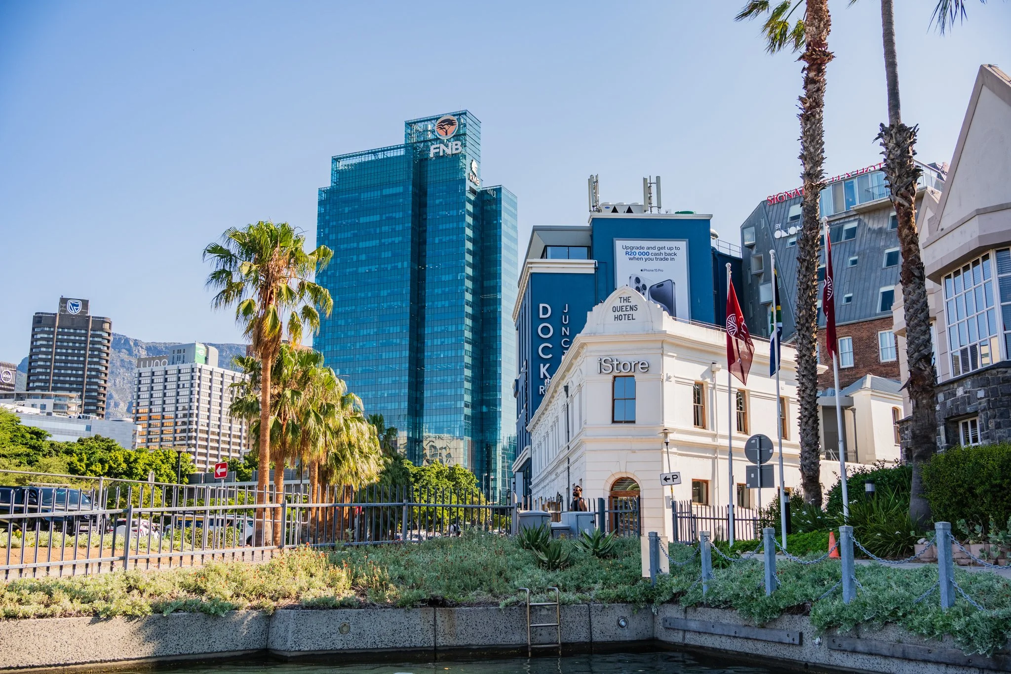 Cityscape with modern glass skyscrapers, palm trees, and historic buildings along a waterway under a clear blue sky.