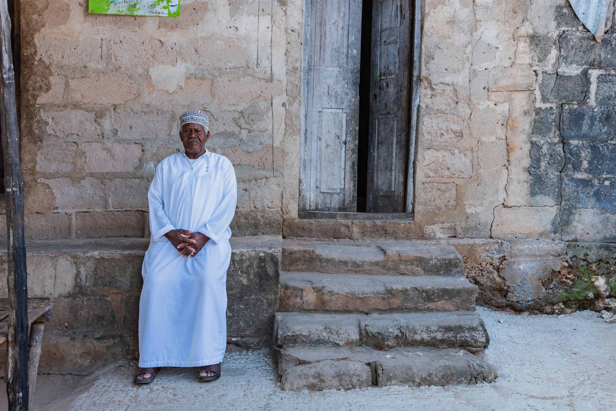 An elderly man in a Kanzu, sitting against a weathered stone wall with a wooden door and steps nearby, outdoors in Zanzibar, Tanzania.