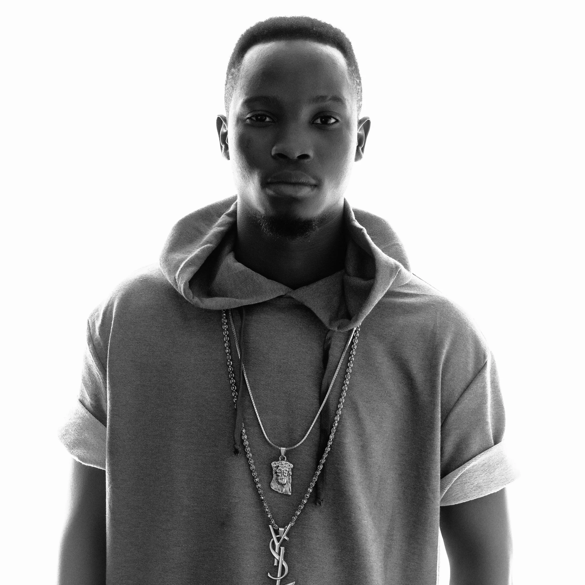 Black and white portrait of a young man with short hair, wearing a hoodie and multiple chains around his neck, standing against a bright background.